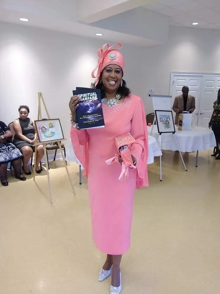 Woman in a pink dress and hat holding a book, smiling at an indoor event.