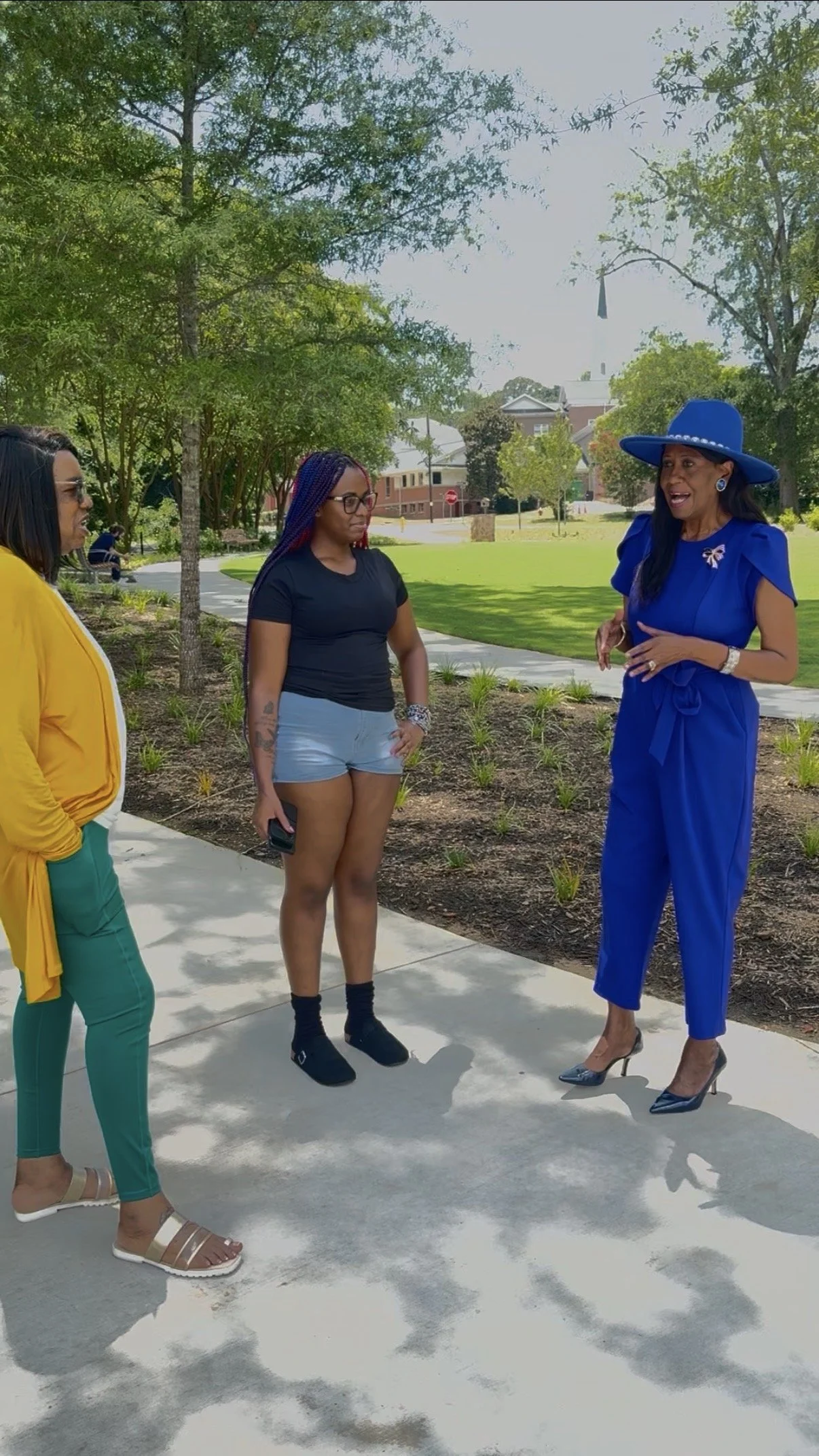 Four women talking outdoors on a sunny day in a park with trees, grass, and a walkway, including one in a blue dress and hat, another in a black t-shirt and denim shorts, one in a yellow jacket and teal pants, and one sitting in the background.