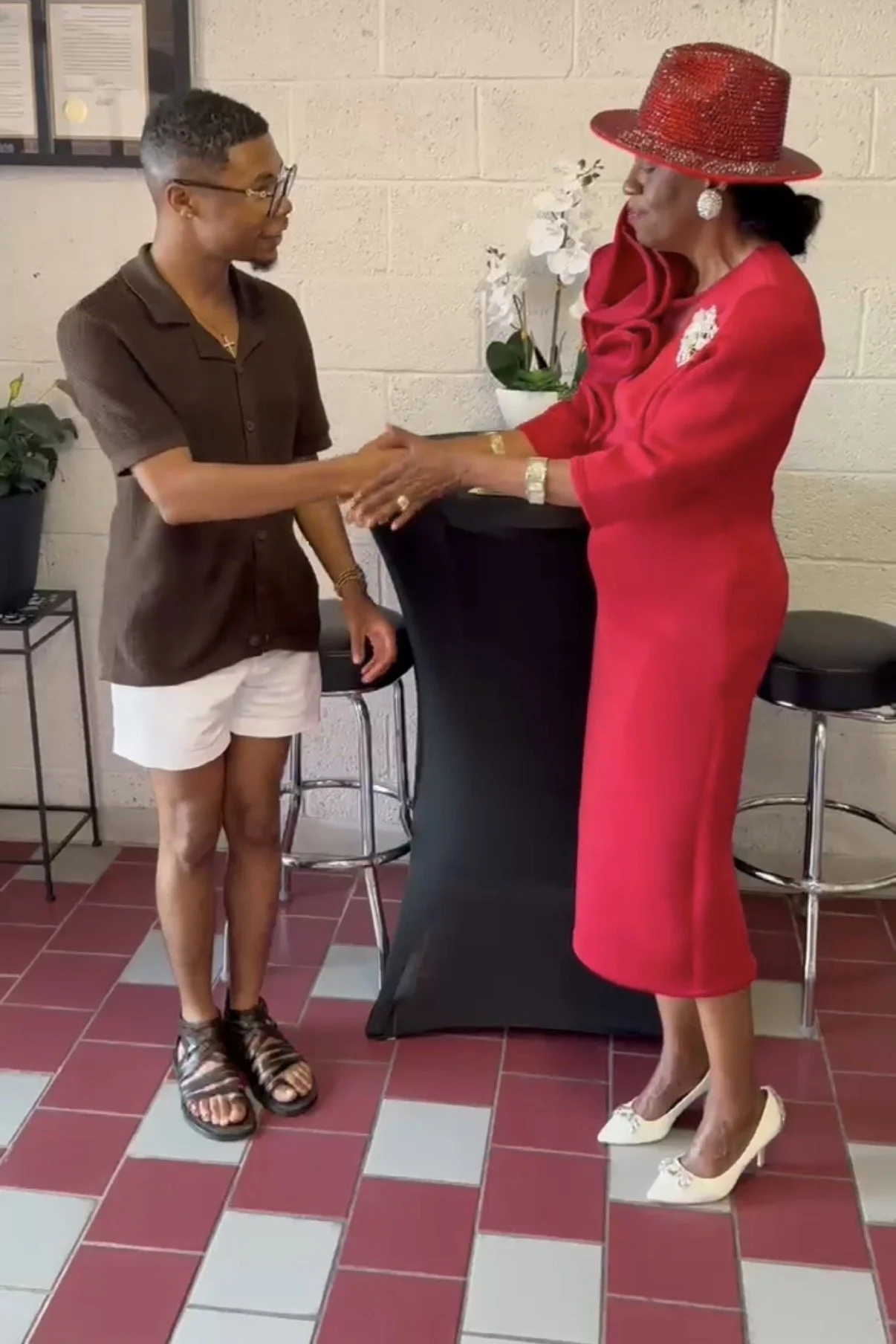 Two women shaking hands indoors, one wearing a brown shirt and white shorts, the other in a red dress and red hat, with potted plants and chairs in the background.