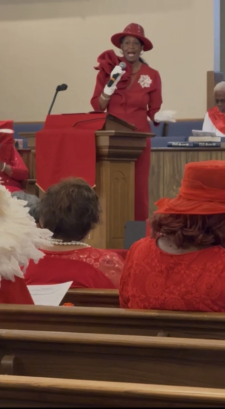 A woman in red and a large red hat speaking at a podium during a service or event, with other women dressed in red seated nearby.