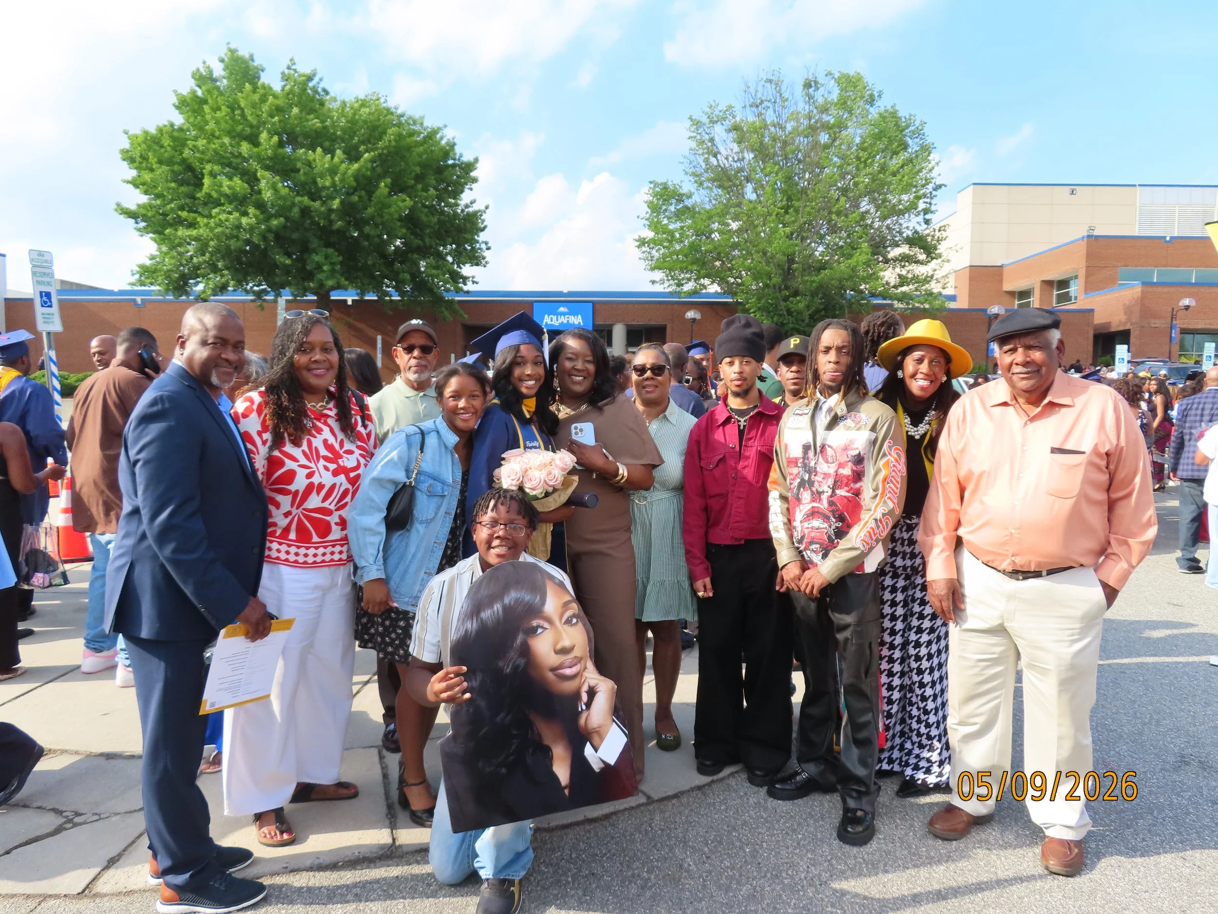 A group of people, including men, women, and children, gathered outdoors in front of a building, commemorating an event. Some are dressed in graduation caps and gowns, indicating a graduation celebration. There are trees and a blue sky in the background. The photo is timestamped May 9, 2026.
