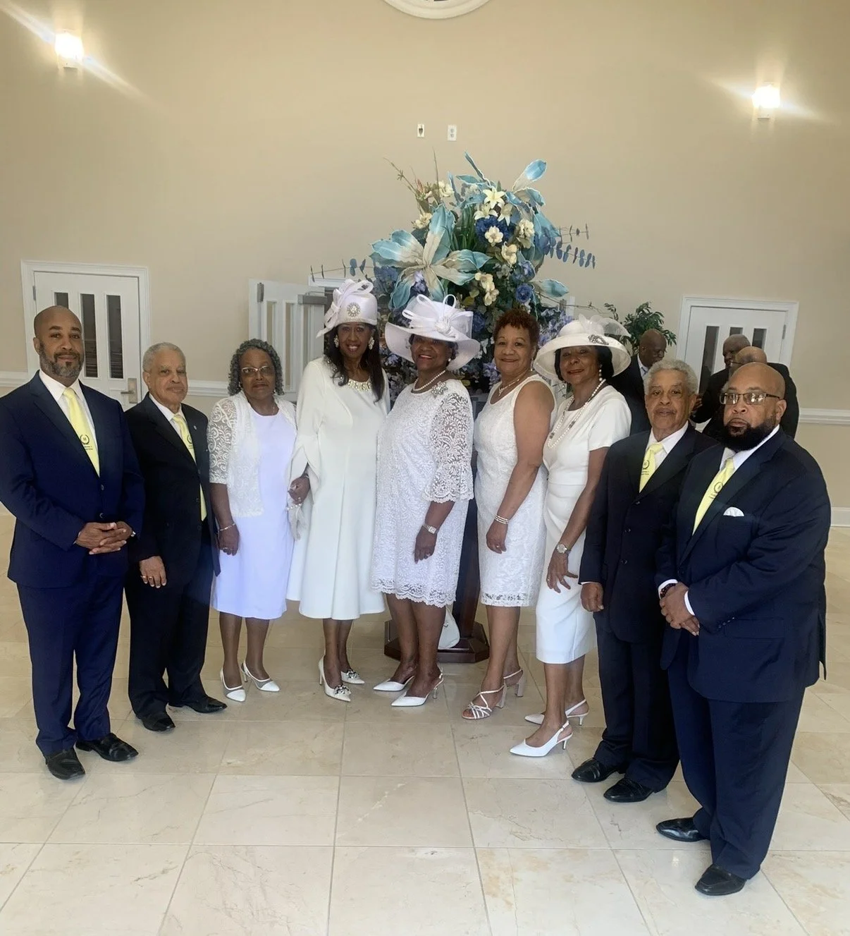 Group of nine people dressed in formal white and navy blue attire, standing in front of a floral arrangement with blue and white flowers at an indoor event or celebration.