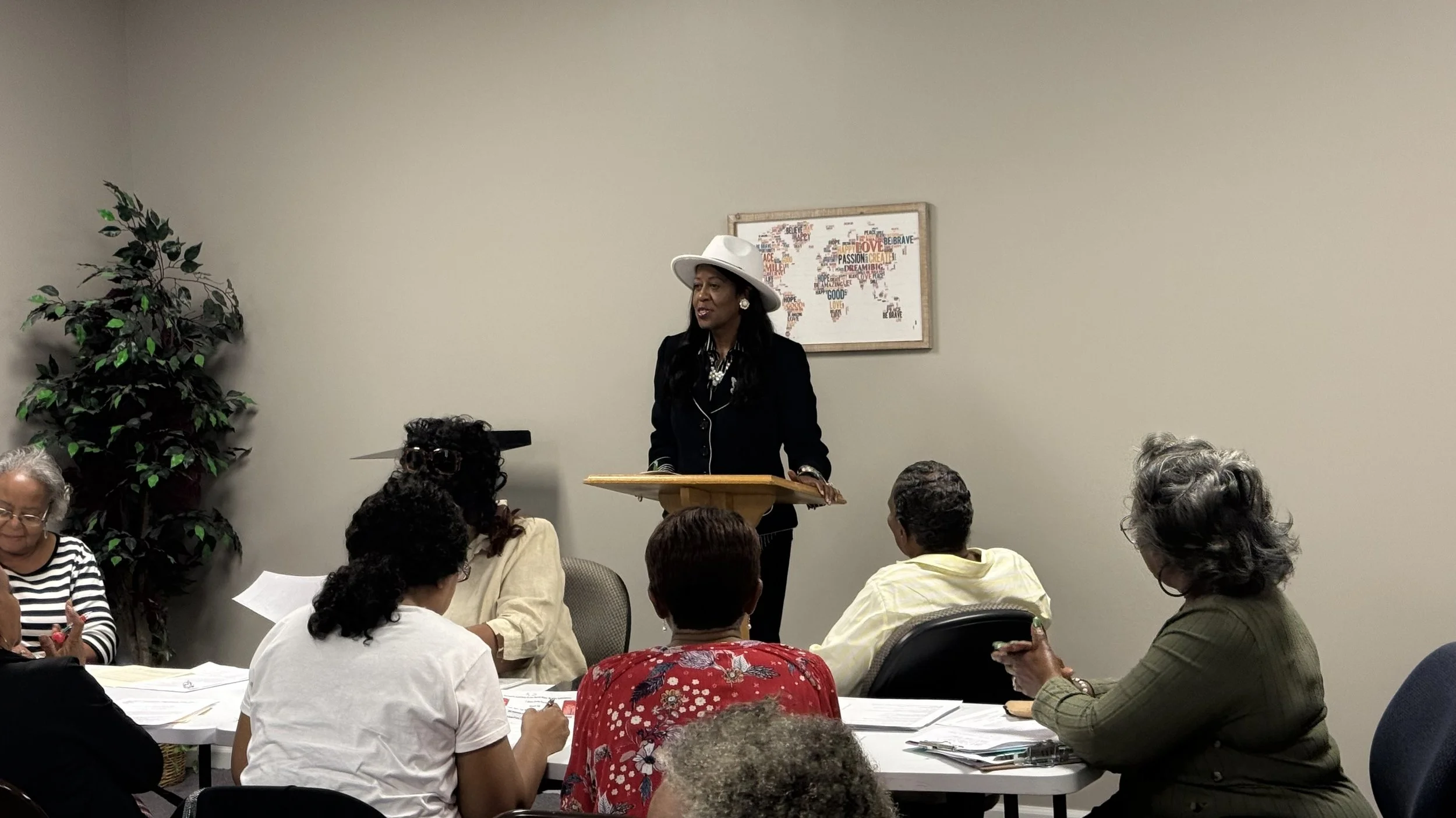 A woman in a white hat and black outfit standing at a podium speaking to a group of diverse adults seated at tables in a meeting room.
