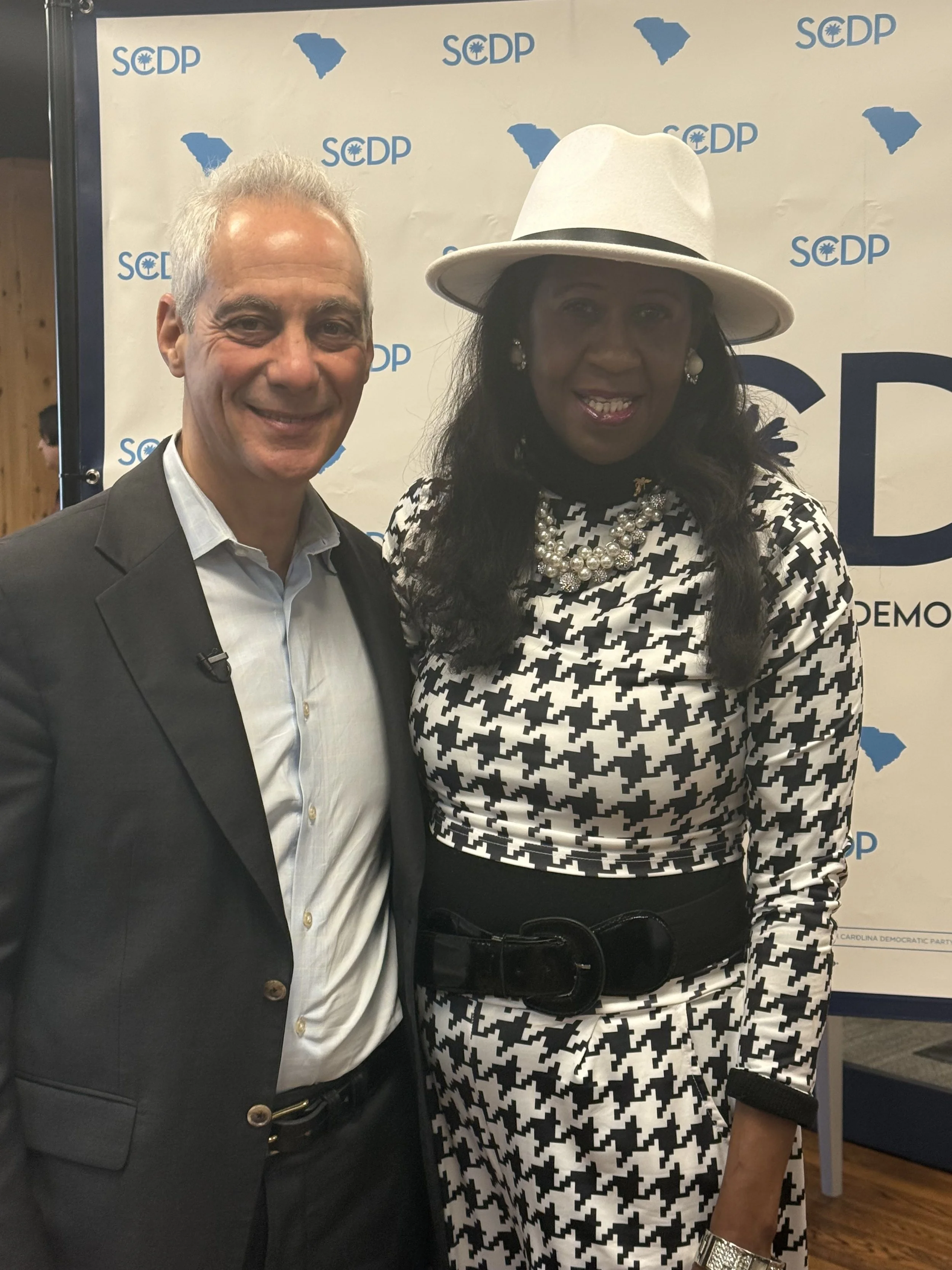 Two people standing in front of a political backdrop with the logo of the South Carolina Democratic Party. One man with gray hair wearing a dark suit and white shirt, and woman wearing a white hat, black-and-white patterned dress, and pearl necklace, smiling at the camera.