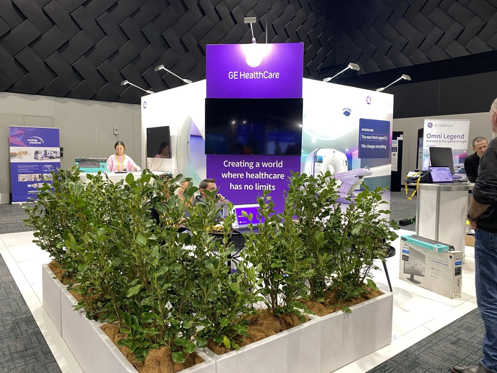 An exhibit booth at a conference or trade show featuring GE Healthcare, including a large screen with the slogan 'Creating a world where healthcare has no limits', various informational displays, and a large image of a medical imaging device. There are people seated and standing around the booth with plants in the foreground.