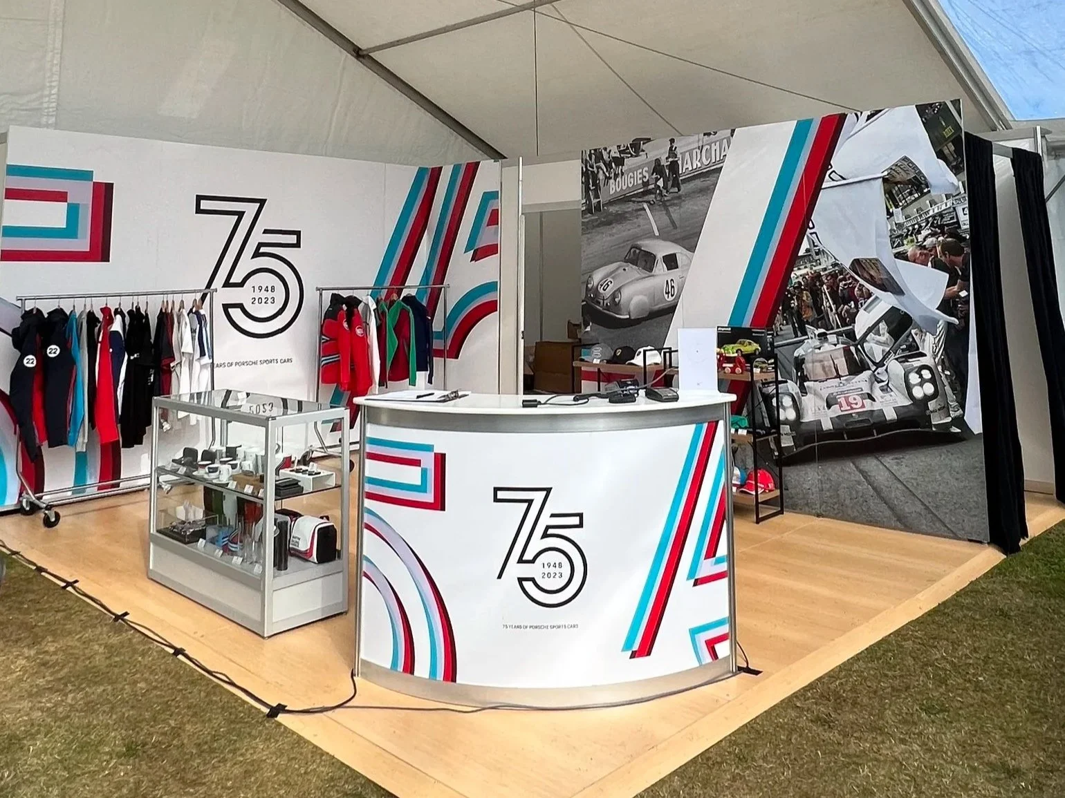 A vintage Porsche display booth celebrating 75 years of Porsche sports cars, with clothing, memorabilia, and black-and-white race car photos.