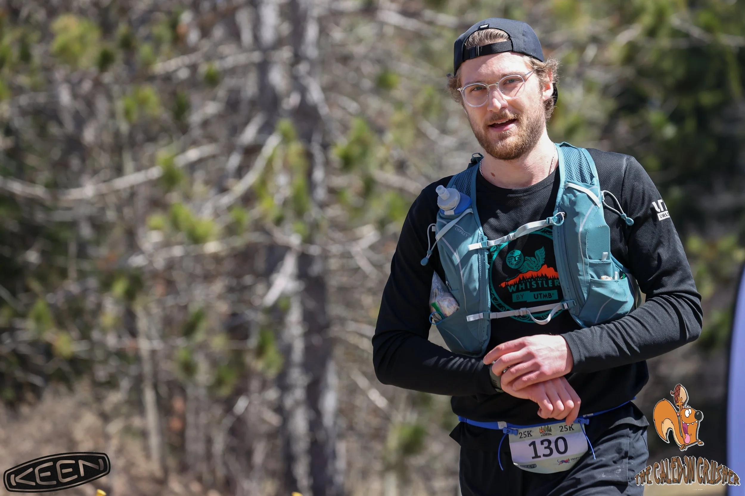 A man with glasses, a beard, and a backwards black cap participating in a race, wearing a blue hydration pack, black long sleeve shirt, and a race bib number 130, with a wooded background.