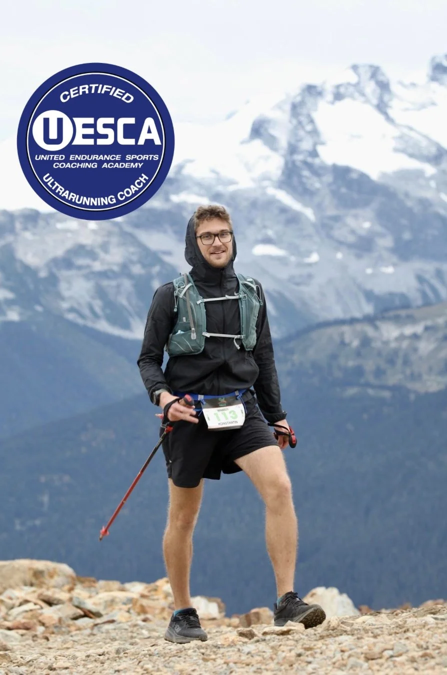 A man in outdoor gear smiling while standing on a rocky trail with mountains in the background during a trail running event, wearing a bib number 13.