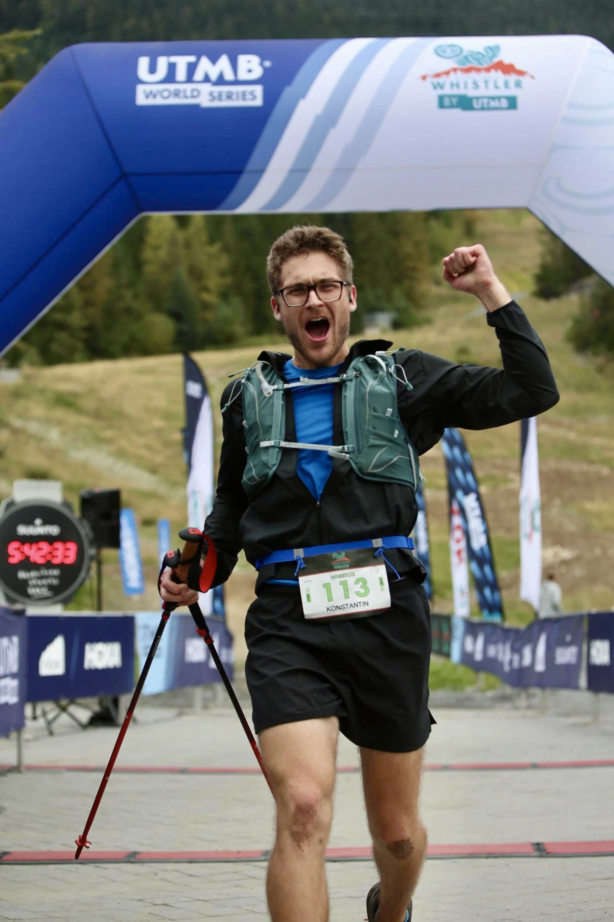 A man with glasses and a beard wears a blue athletic shirt, black shorts, and a backpack, running in a race with a bib number 113, crossing the finish line with his right arm raised in celebration. The background shows a mountainous area with trees, banners, and a digital timer showing 5:42:33.
