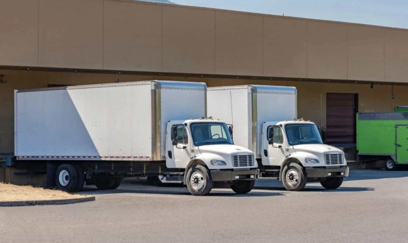 Two white box trucks parked side by side in front of a beige building with a brown door and a green trailer to the right.