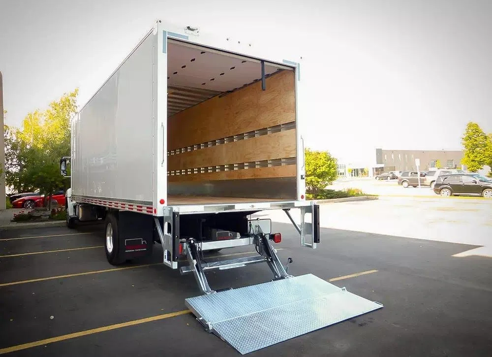 Empty moving truck with a lowered wheelchair ramp in a parking lot with some parked cars and trees in the background.