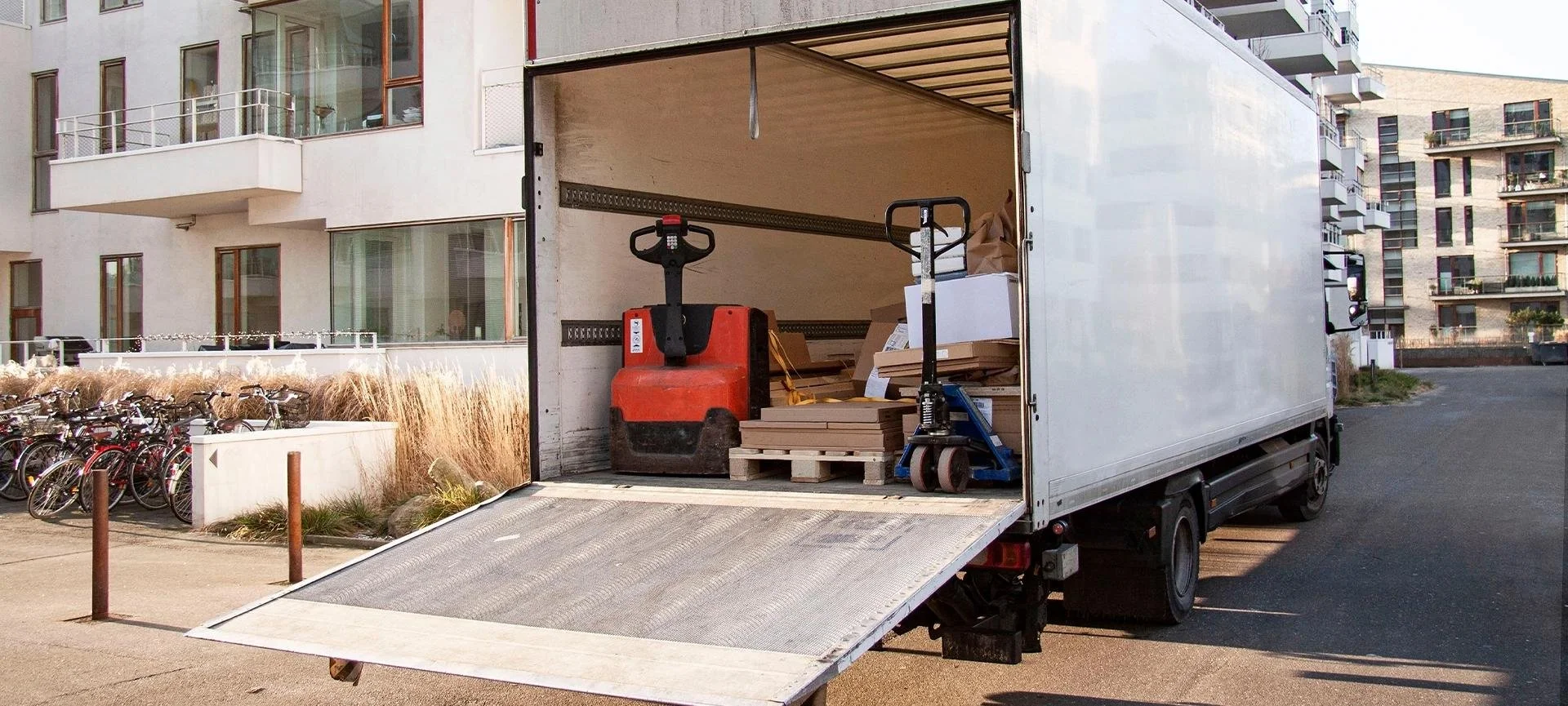 Open white moving truck with wooden pallets, cardboard boxes, and a pallet jack inside, parked in front of a modern apartment building.