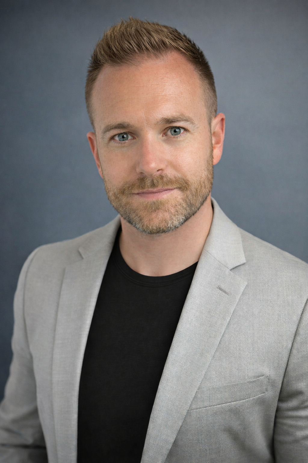 Headshot of a man with short, styled blond hair, blue eyes, and a beard, wearing a light gray blazer over a black shirt, against a dark gray background.