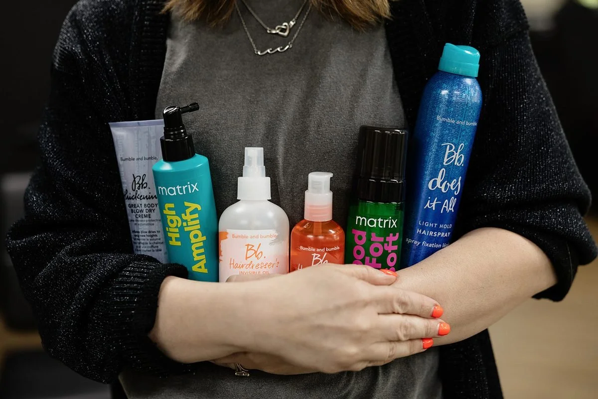 An armful of shampoo, styling spray and hair oil bottles held at WAREHAUS Hair Studio, the kind of product layering that leads to scalp buildup