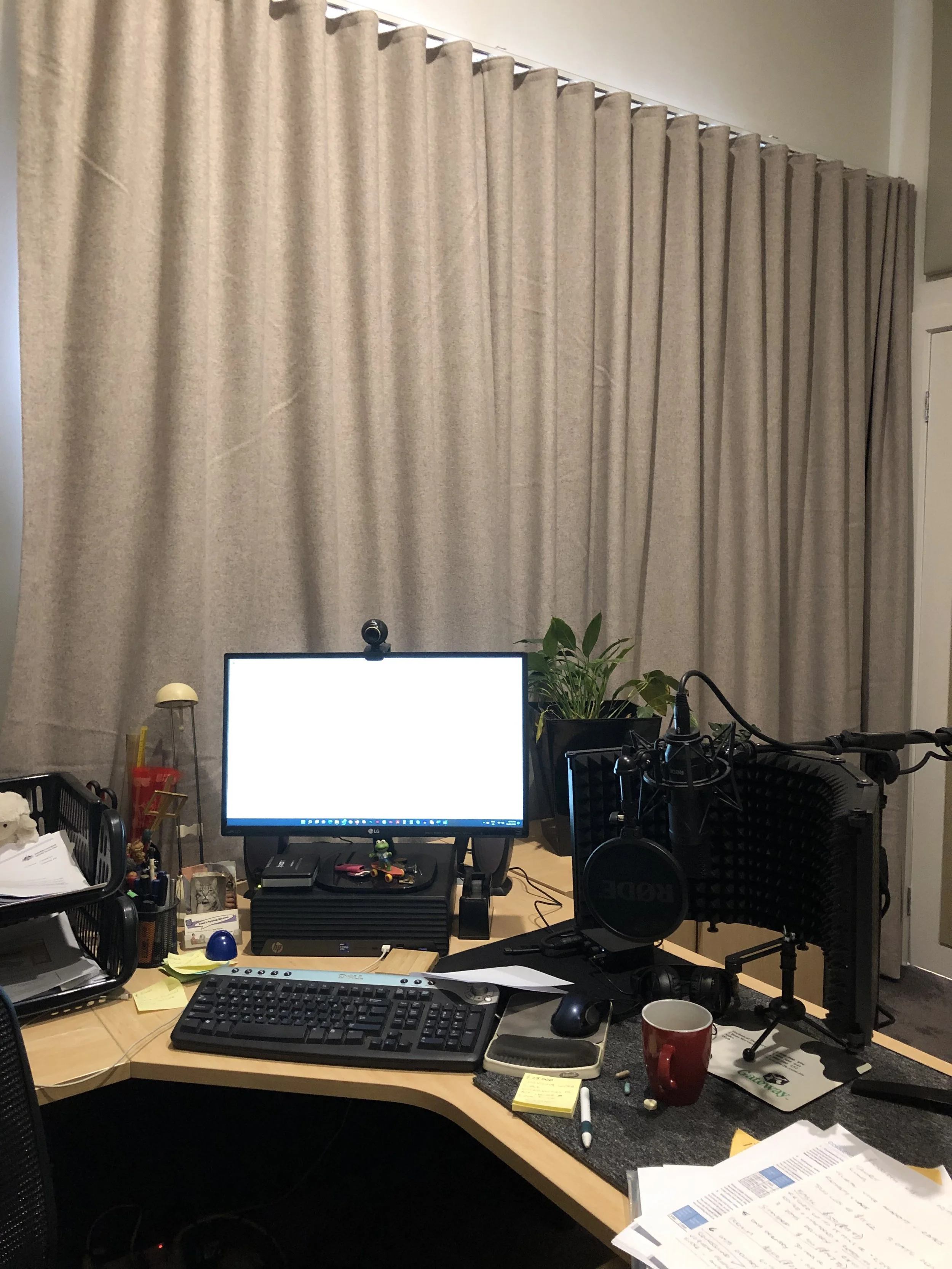 A cluttered office desk with a computer monitor, keyboard, set of papers, coffee mug, and various office supplies, with beige curtains in the background.