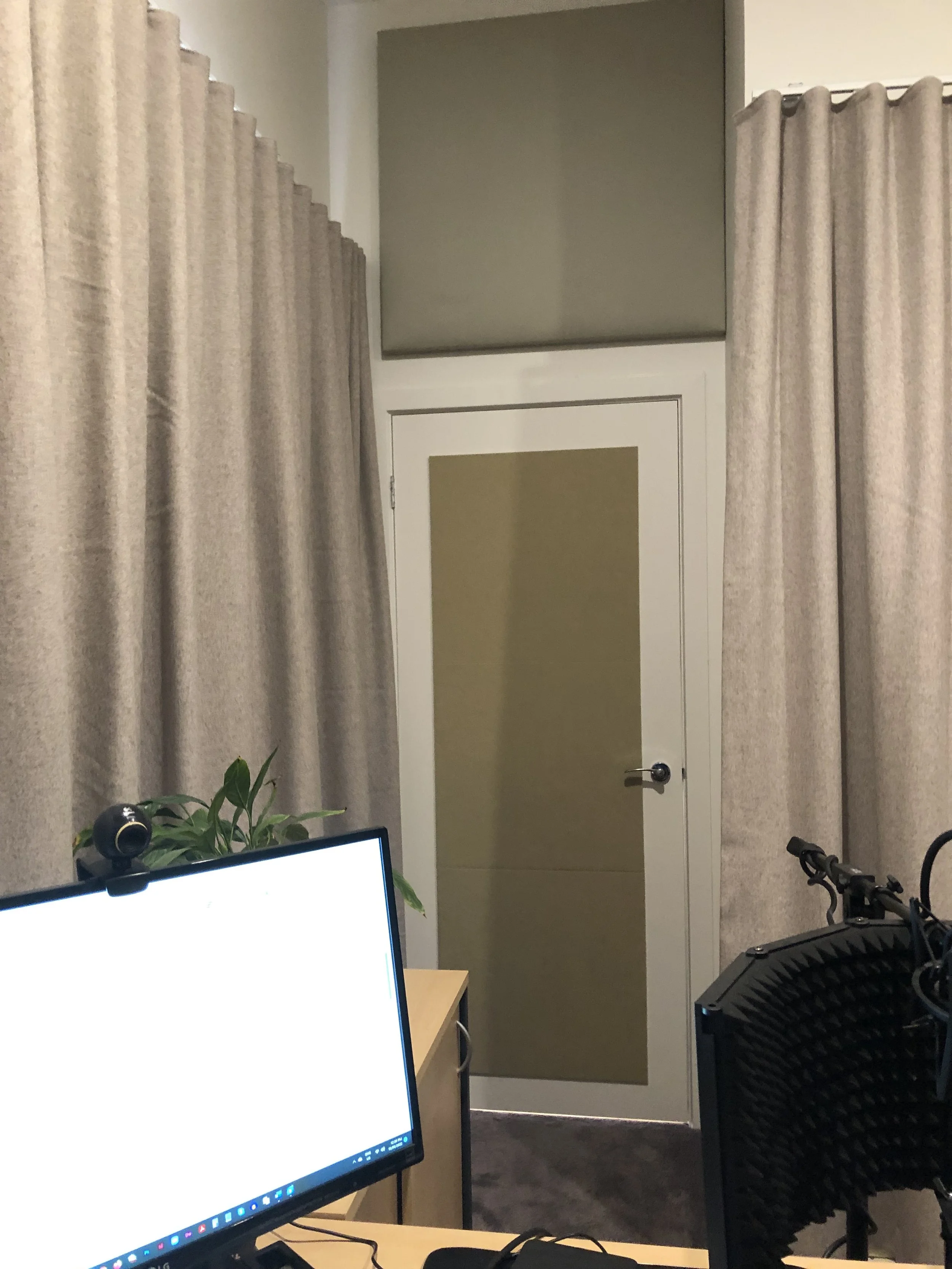 A corner of an office with beige curtains, a door with frosted glass, and a computer monitor on a wooden desk. There are some plants and equipment visible around.