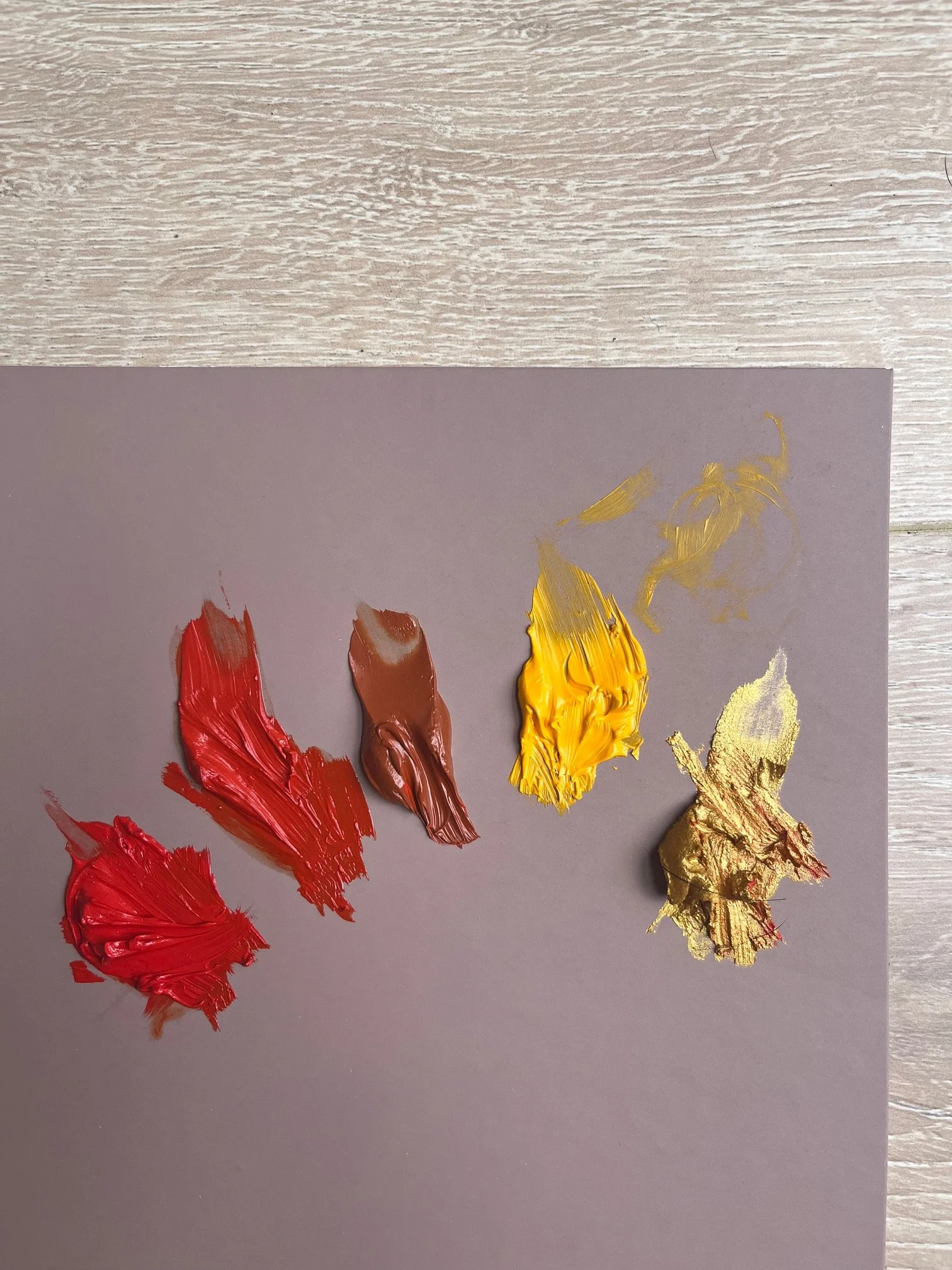 Close-up of a gray palette with thick, textured strokes of red, brown, yellow, and gold paint on a gray background, with a wooden surface underneath.