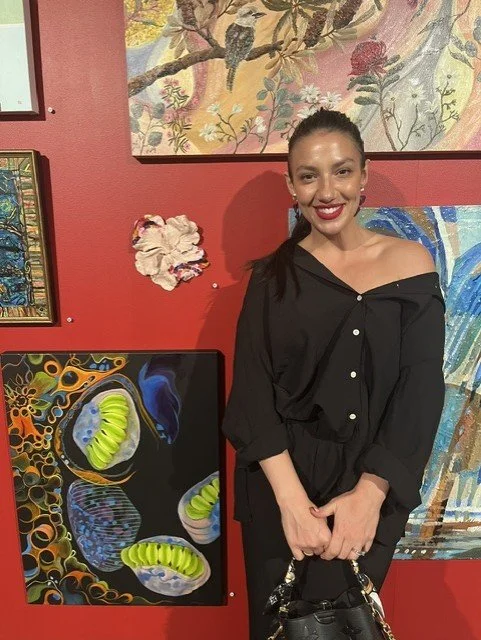 A woman with dark hair, tied back, wearing a black off-the-shoulder blouse and smiling at the camera, standing in front of colorful artwork in a gallery or art display setting.