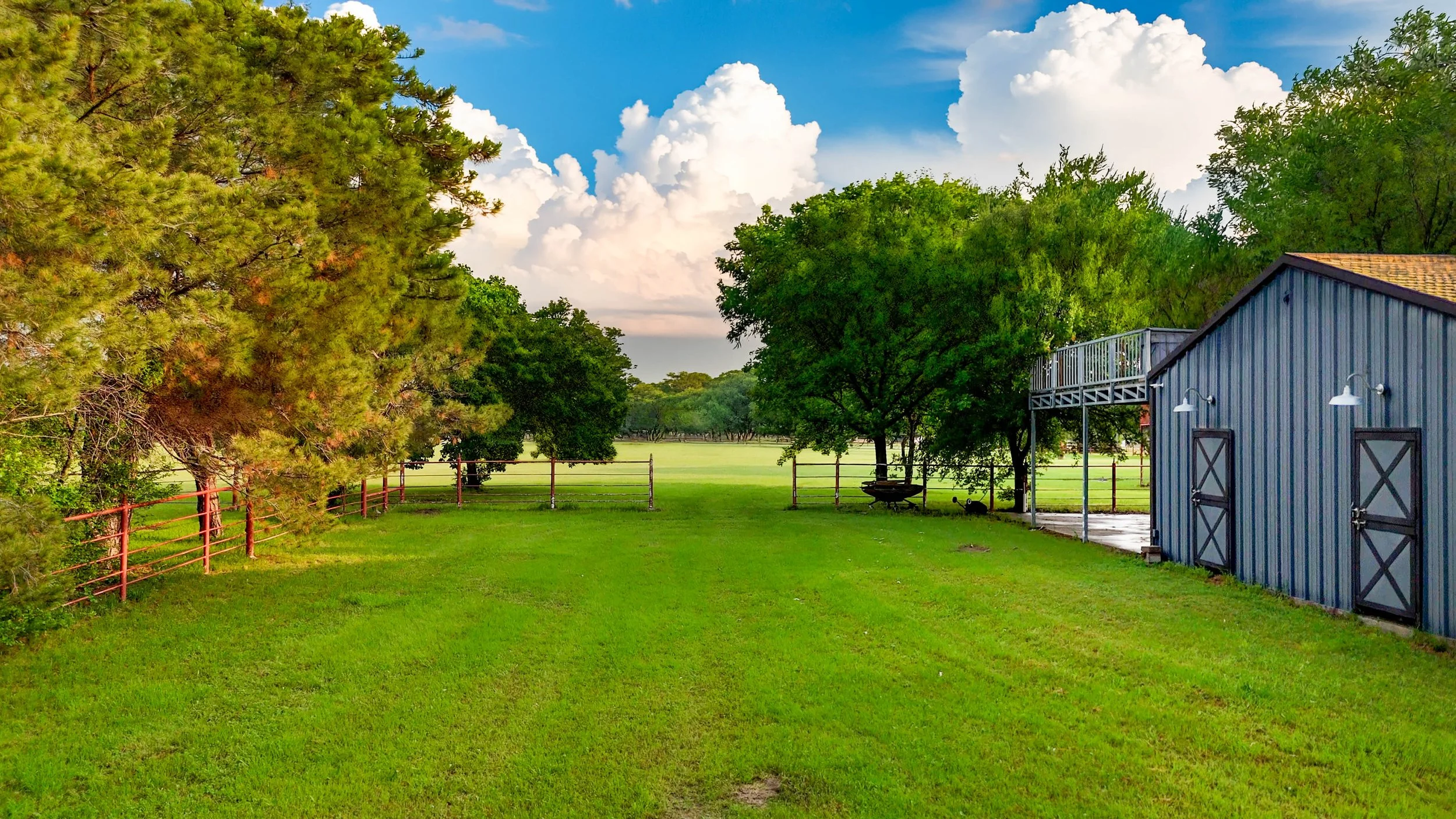 A lush green backyard with a red fence, tall trees, a blue barn with white doors, and a cloudy sky overhead.