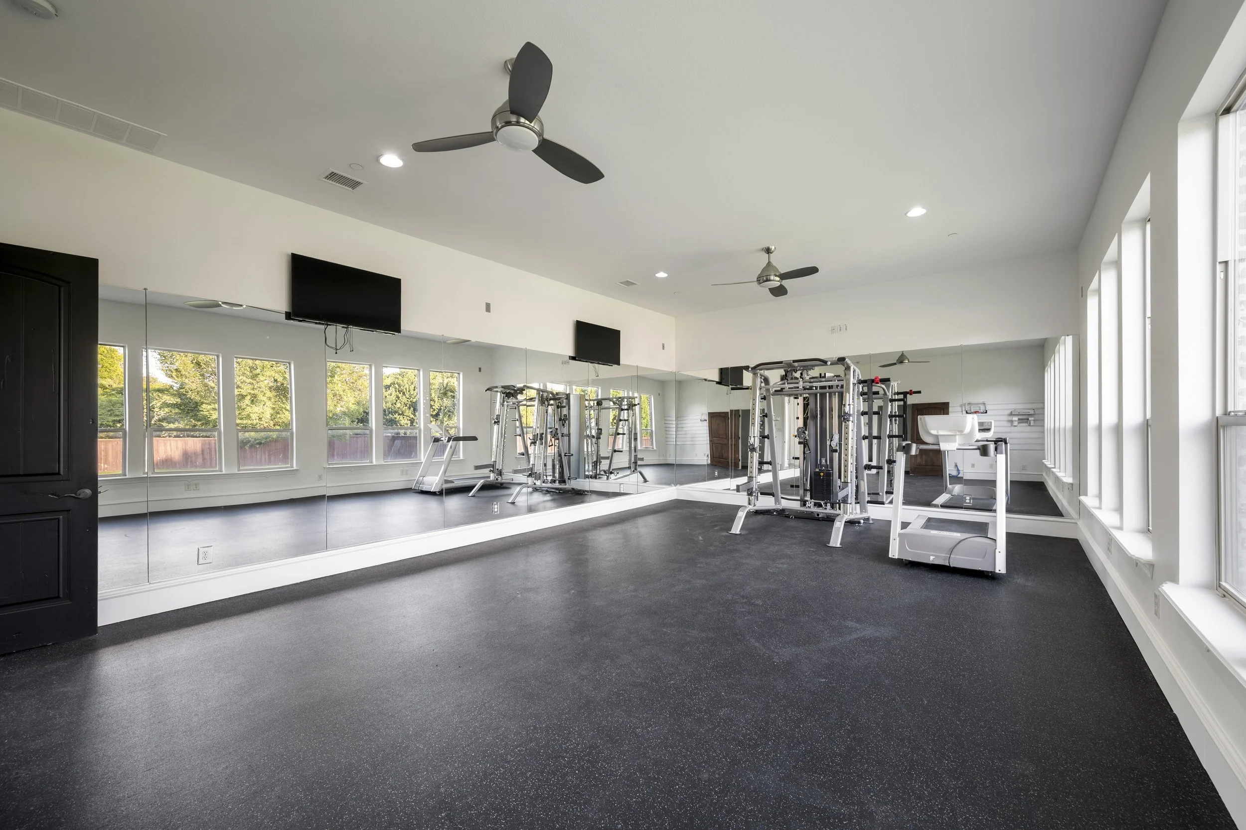 A spacious gym with large windows, black rubber flooring, and fitness equipment including weight machines and a treadmill, with ceiling fans and wall-mounted TVs.