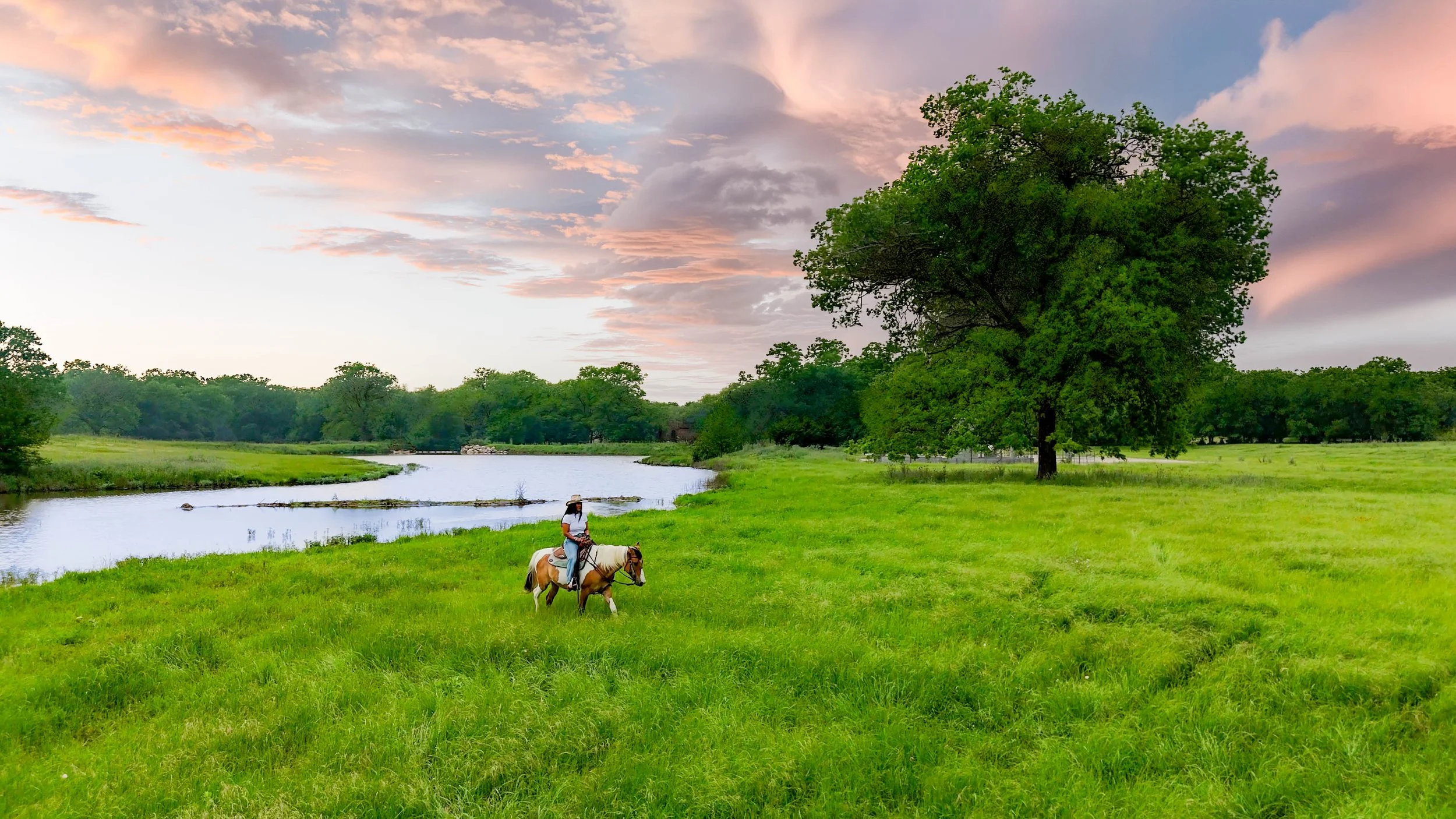 A person riding a horse across a lush green field near a river with trees, under a sky with pink and purple clouds at sunset.