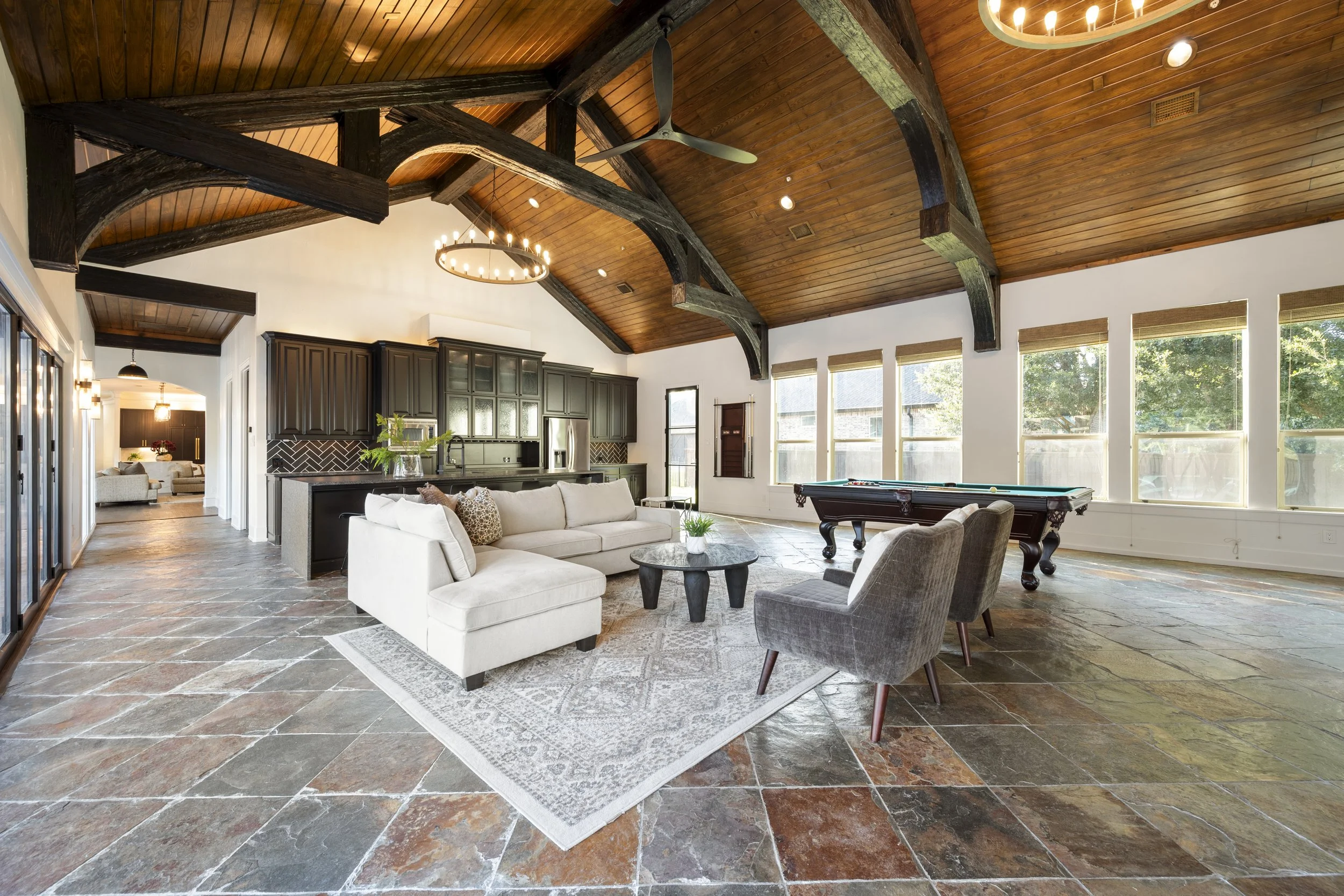 Living room with a high wooden ceiling featuring exposed dark wood beams and a ceiling fan. Large windows allow natural light and offer a view of trees outside. The room has a white sofa, a gray armchair, a small round coffee table with plants, and a