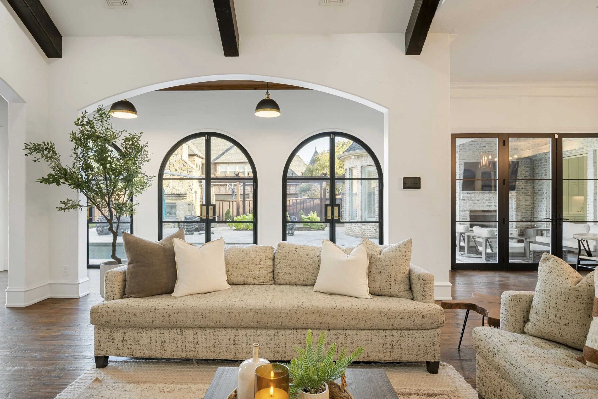 Living room with beige sofa and throw pillows, large arched windows showing an outdoor patio area, a potted plant, and ceiling beams.