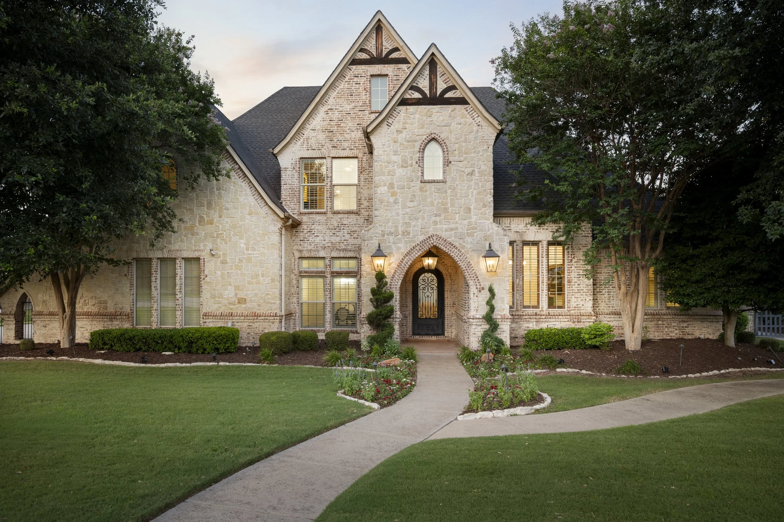 A large, elegant two-story house with a stone and brick facade, surrounded by a well-maintained lawn and lush trees, with a curved pathway leading to the front door.