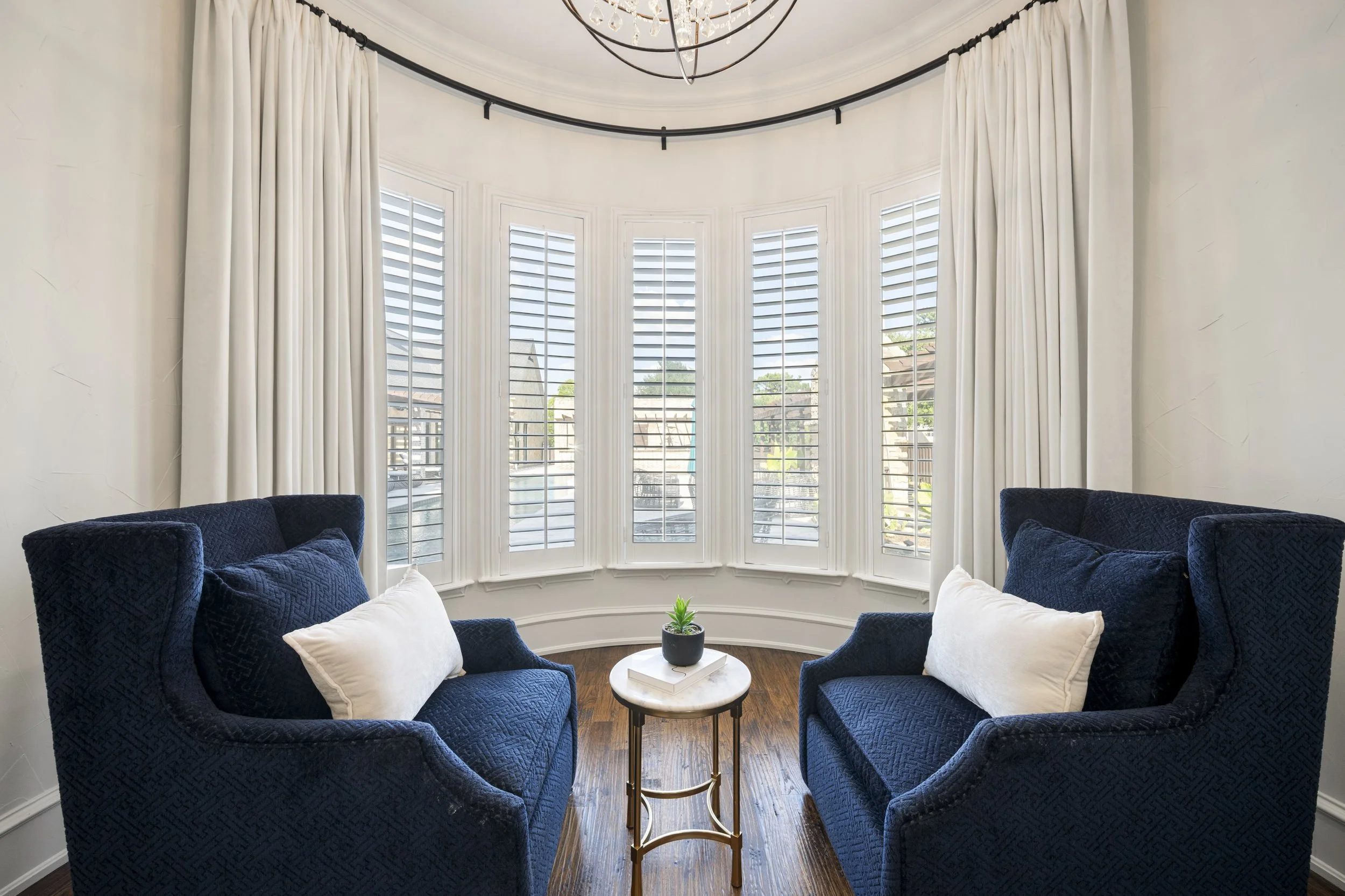 Living room with a bay window, white curtains, blue armchairs with white pillows, a small round table with a small potted plant, and a chandelier hanging from the ceiling.
