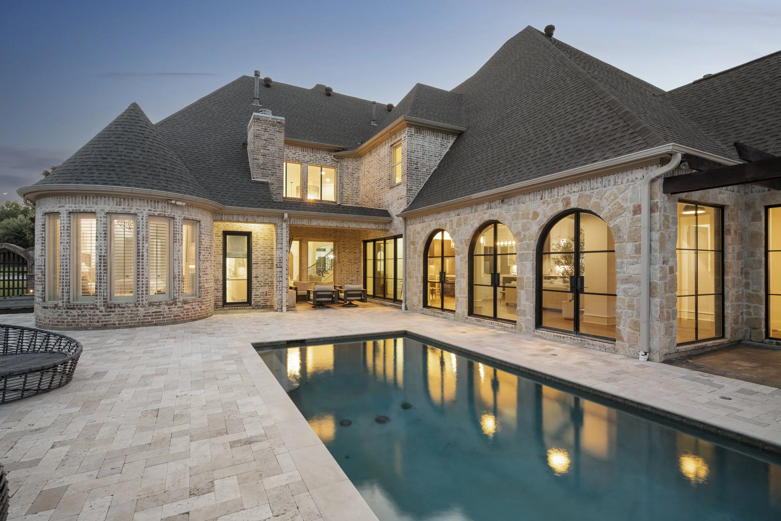 Backyard view of a large brick and stone house with a swimming pool, patio furniture, and illuminated interior at dusk.