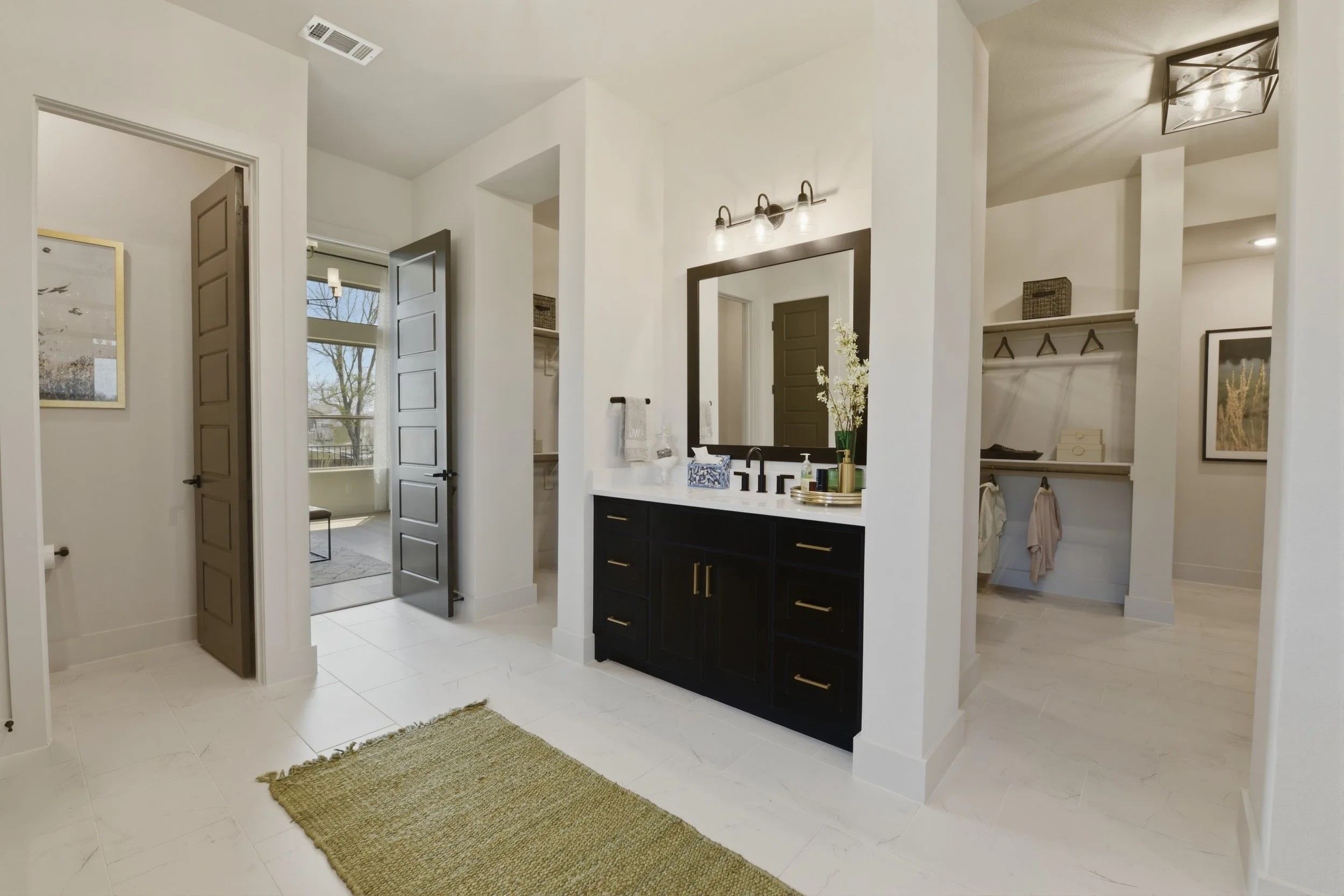 Modern bathroom with black vanity, large mirror, white walls, open closet with hanging clothes, window, and decorative items.