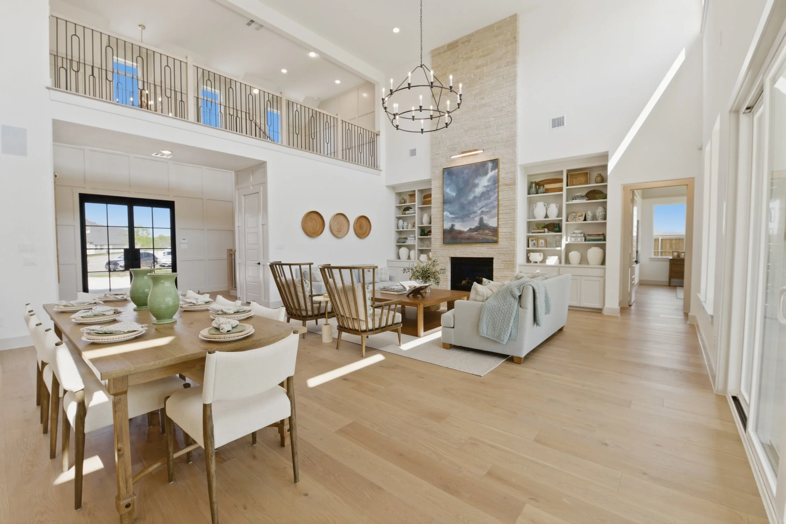 Open-concept living and dining room with a high vaulted ceiling, fireplace, open shelving, chandelier, and large windows letting in natural light.