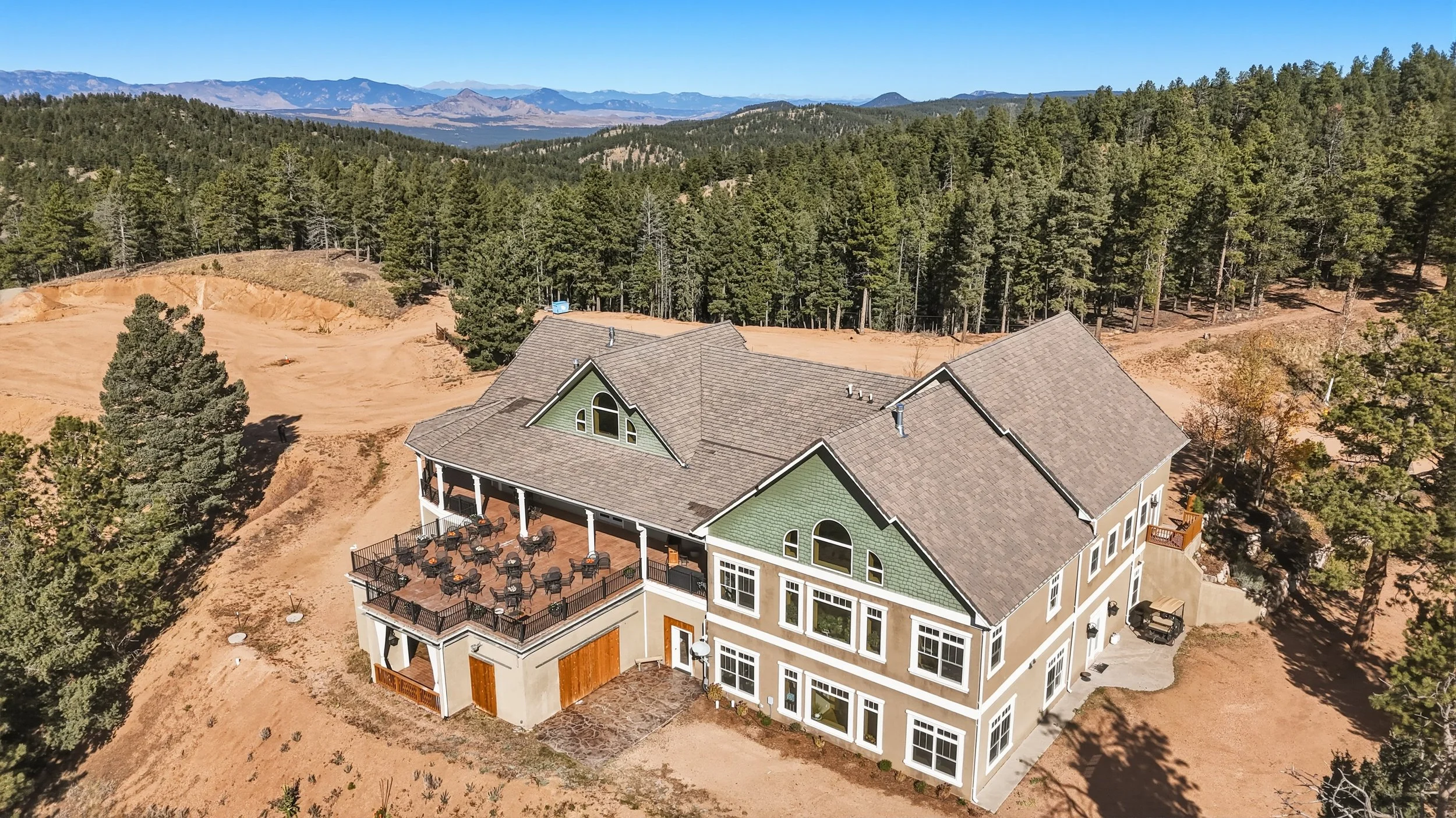 A large multi-story house with a green exterior and numerous windows, featuring a spacious balcony with outdoor seating, situated in a wooded, mountainous area with mountain ranges in the distance under a clear blue sky.
