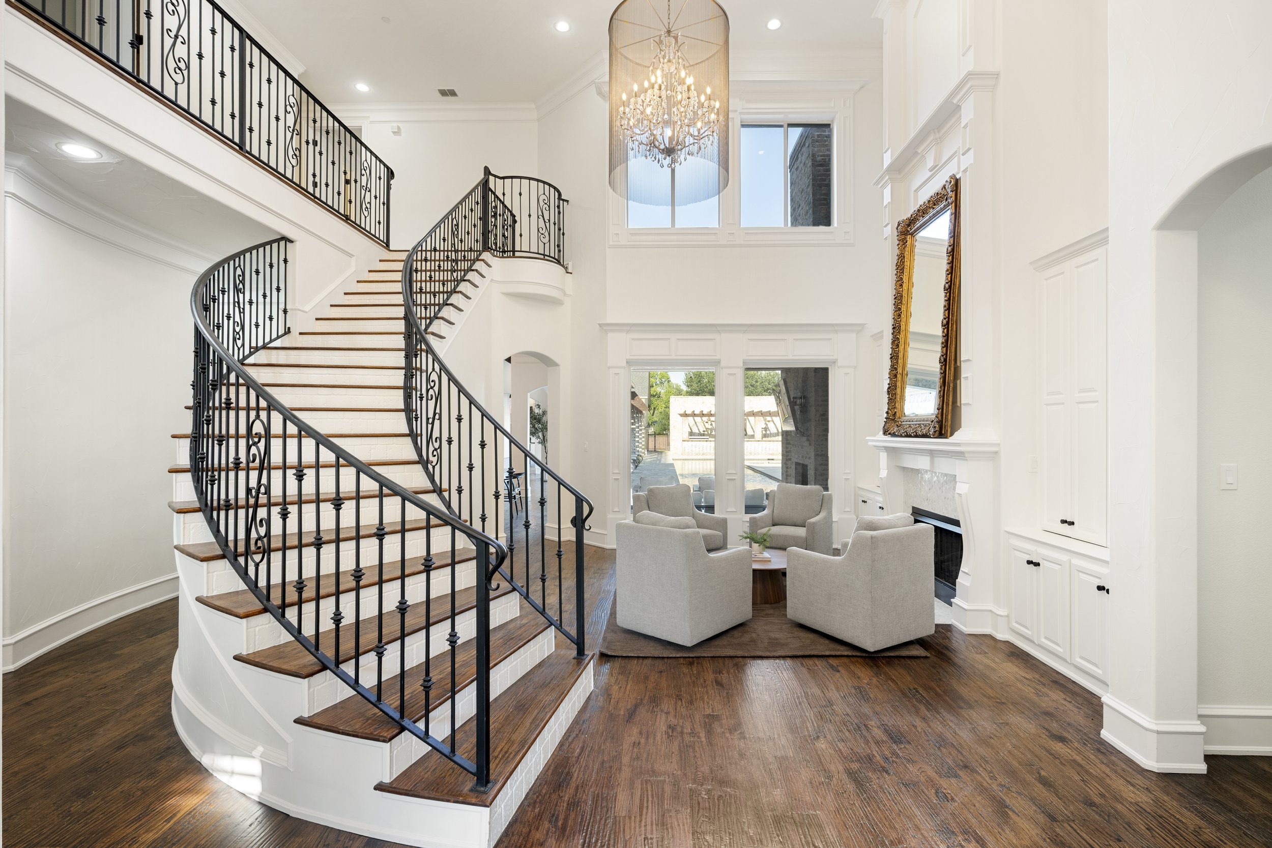 Interior of a spacious, bright living area with a grand staircase featuring black iron railings and wooden steps, cream-colored walls, a large window with a view of greenery outside, a chandelier hanging from the ceiling, a fireplace with a gold-fram