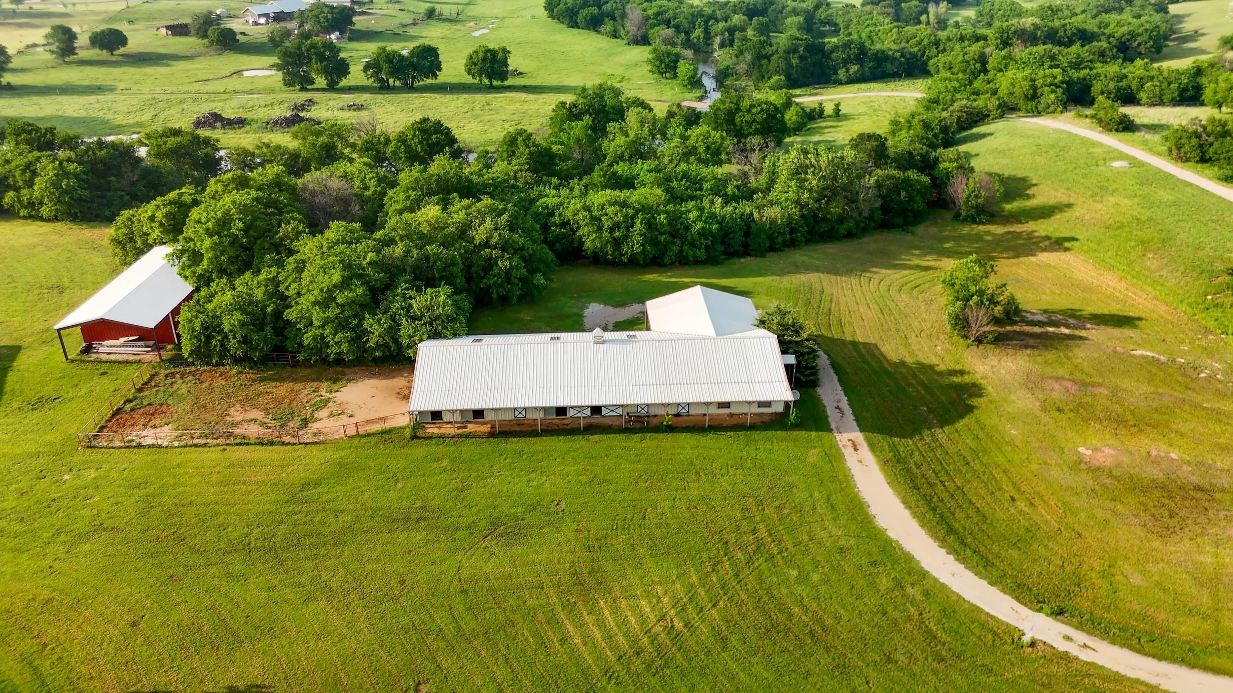 Aerial view of a rural farm with a large white barn, a smaller red barn, surrounded by green trees and open fields, with a gravel driveway leading to the buildings.