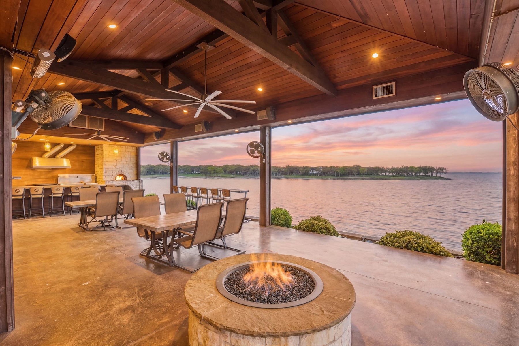 Indoor patio with wooden ceiling, ceiling fans, and outdoor fireplace overlooking a lake at sunset. Furniture includes tables and chairs, and a fire pit is in the foreground.