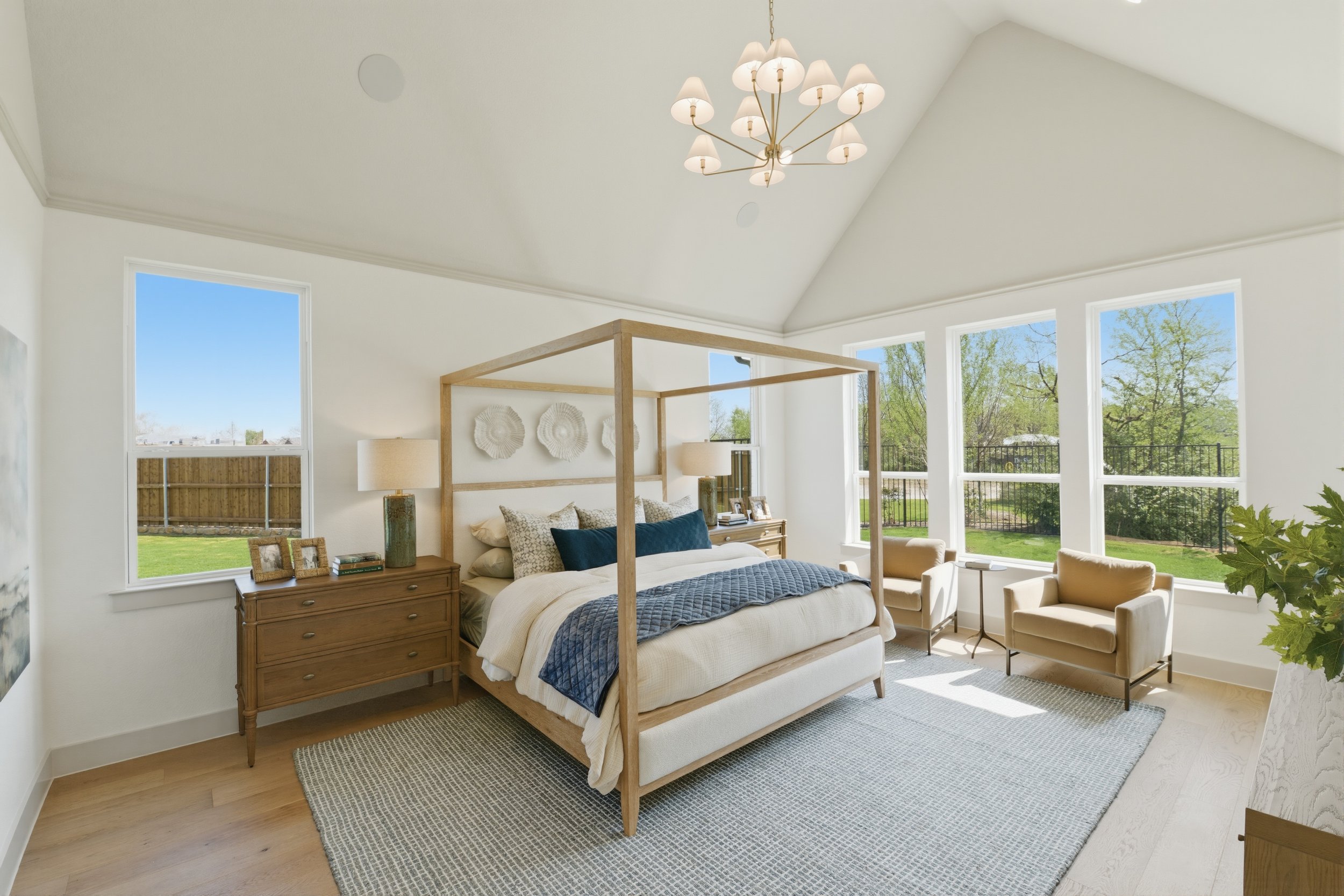Bright bedroom with a white bed, wooden canopy frame, beige armchairs, wooden nightstand, and large windows showing green trees outside.