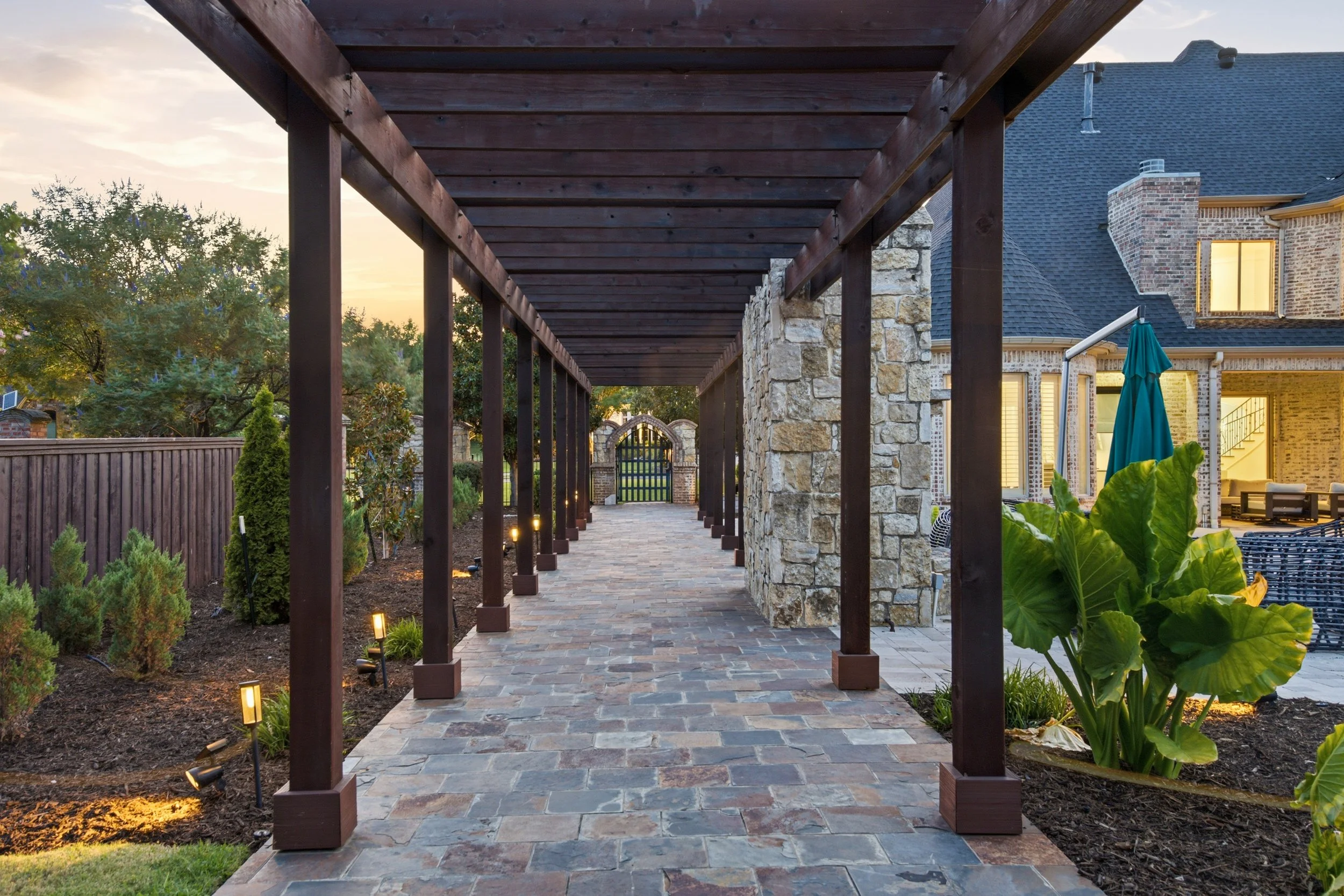 A covered outdoor walkway with a brick floor, supported by dark wooden beams, leading to a stone gate with a wrought iron door. On either side, there are plants and outdoor lighting.