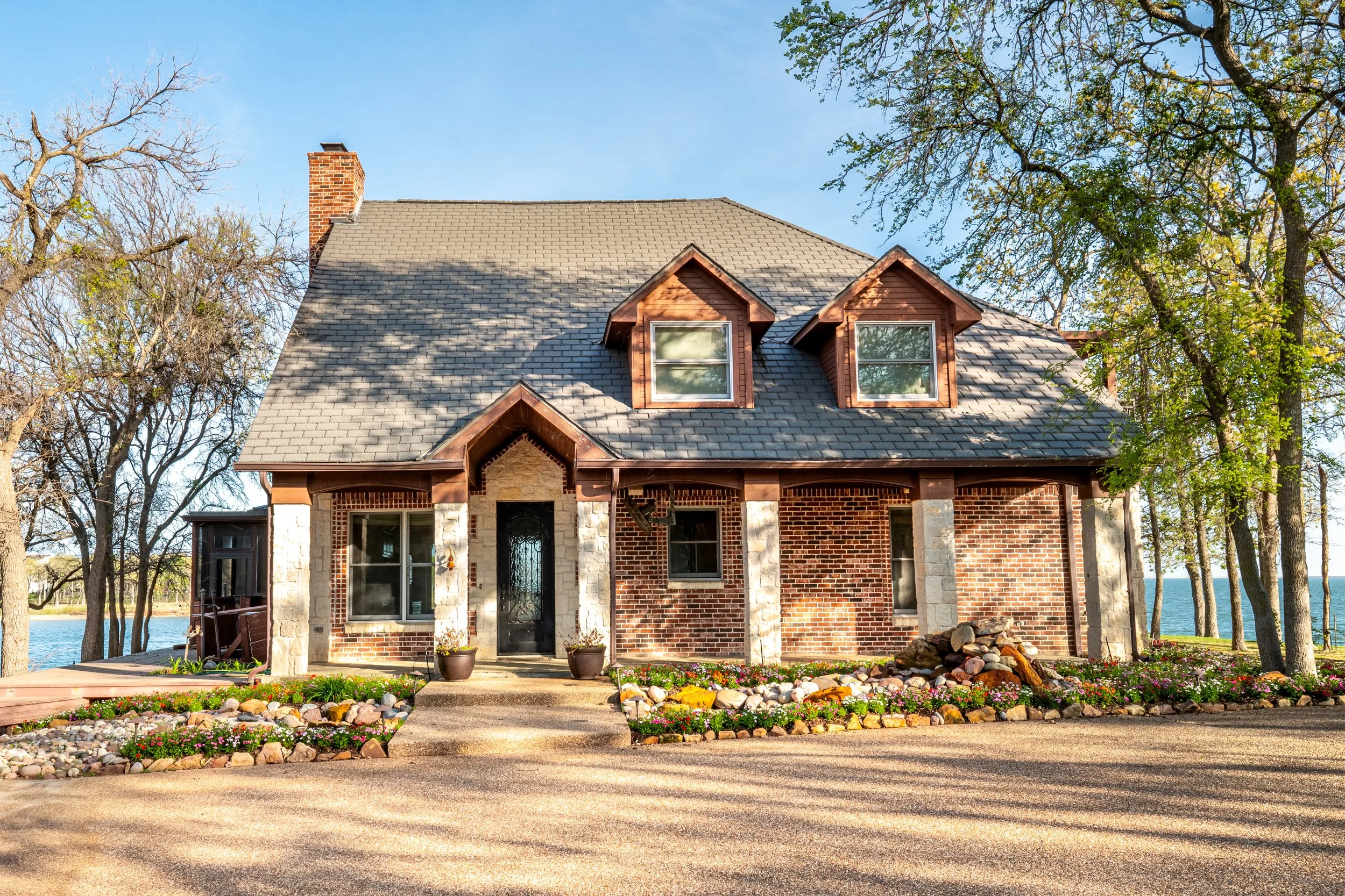 A two-story brick house with a stone front porch, gabled roof with three dormer windows, and a chimney. The house is surrounded by trees and a landscaped garden with flowers and rocks in the front yard. There is a body of water visible behind the hou