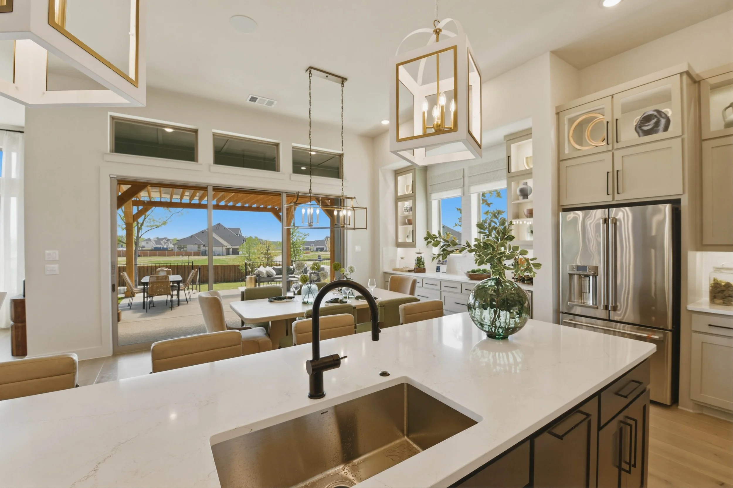 Open-concept kitchen and dining area with white cabinets, a large island with a white marble countertop, and modern light fixtures. Large windows and sliding glass doors let in natural light, showing a backyard with patio furniture and a pergola.