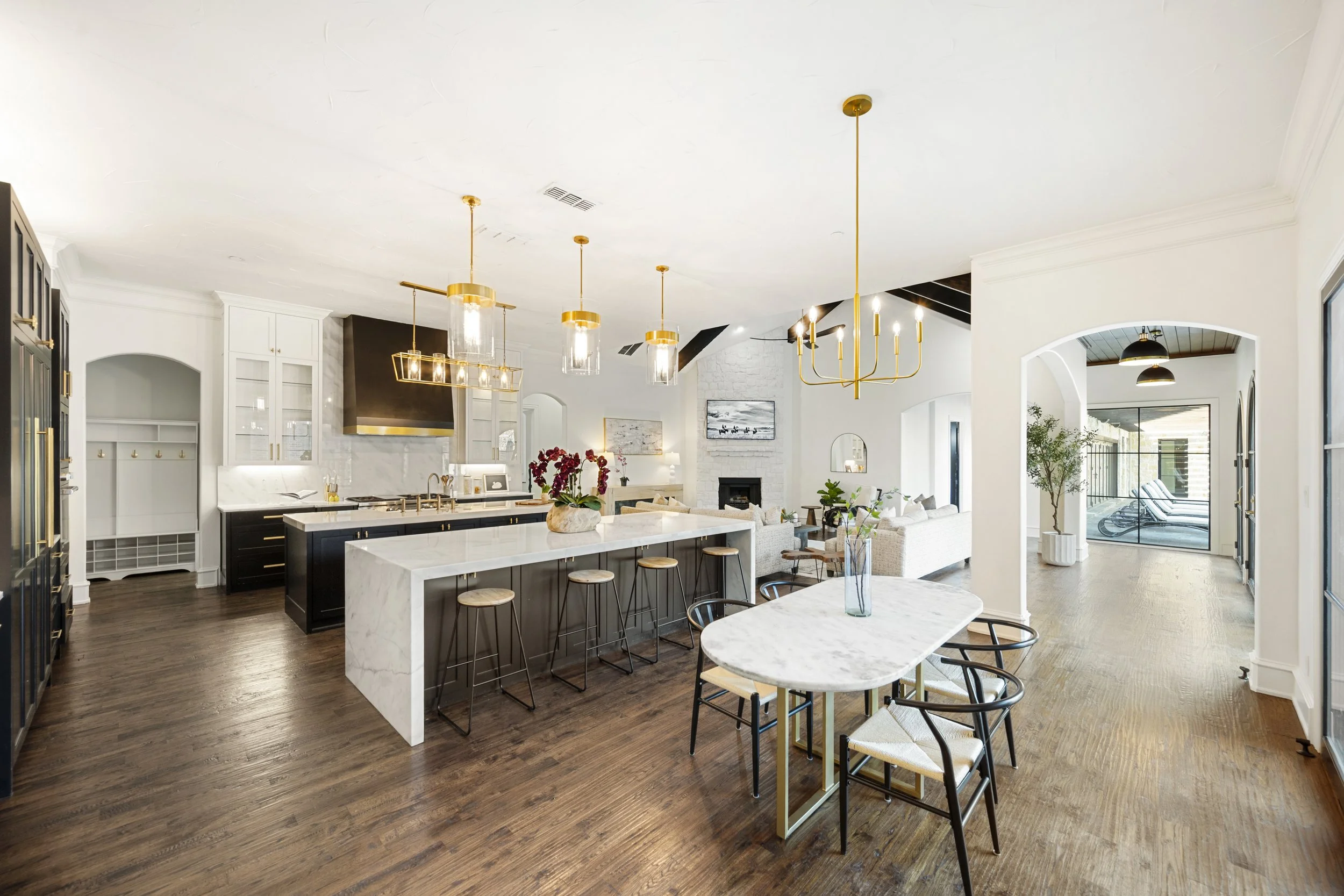 Open-concept modern kitchen and living area with chandelier lighting, white cabinetry, a black and gold kitchen island, and a seating area with sofa and chairs, hardwood floors, large windows, and a balcony.