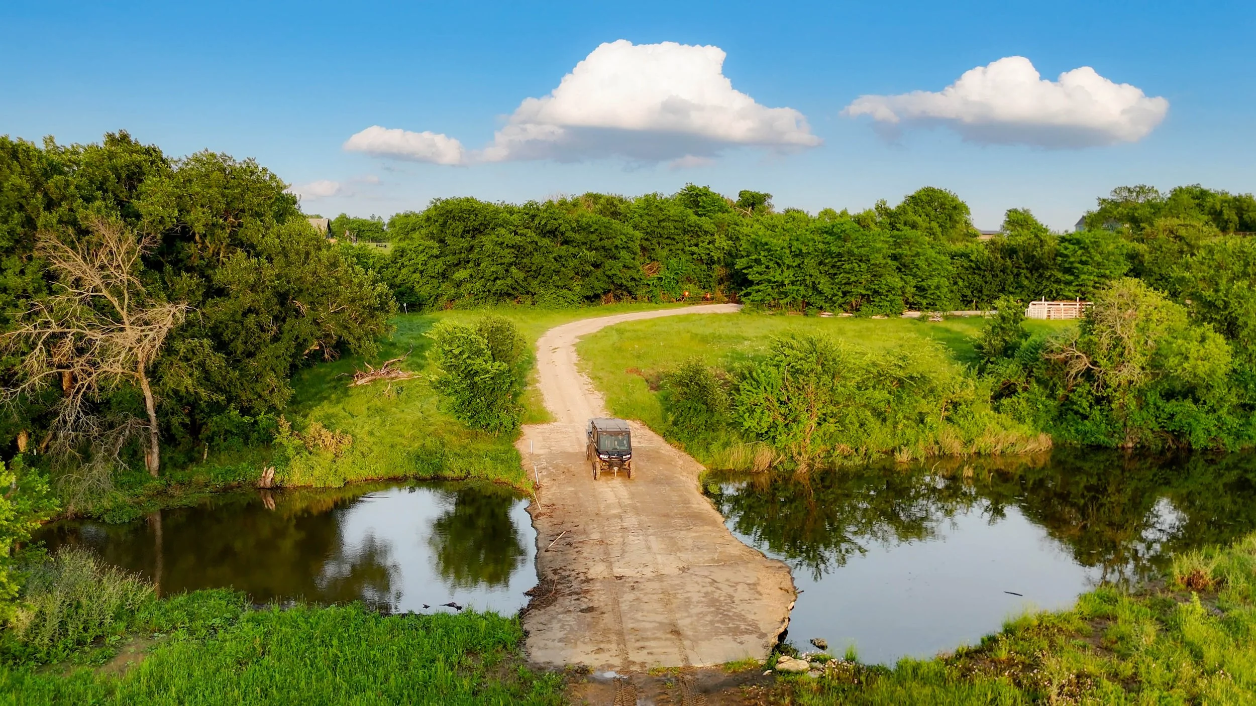 A dirt bridge over a calm river with a vehicle crossing, lush green trees on both sides, and a blue sky with white clouds above.