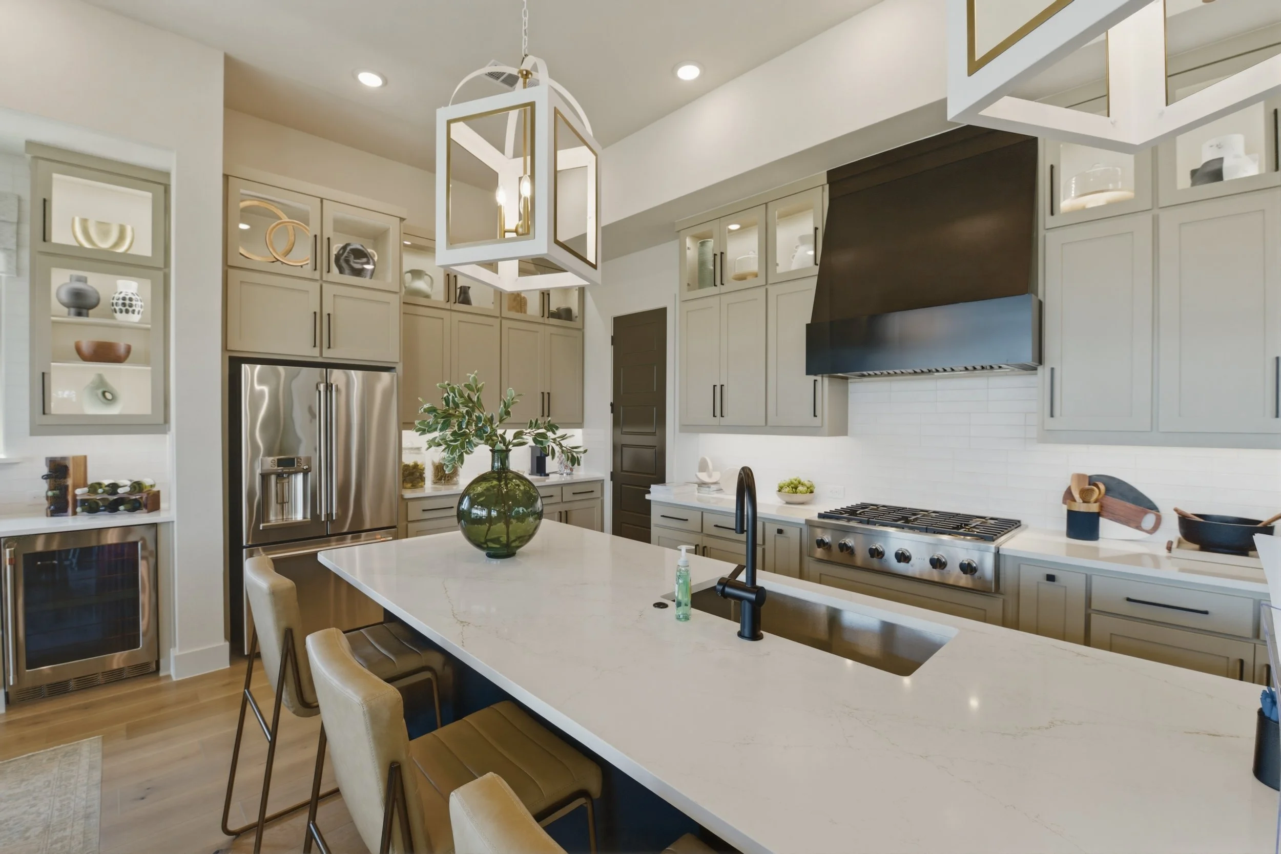 Modern kitchen with white cabinets, stainless steel refrigerator, black range, and white stone island counter with beige chairs. Decorative items displayed in glass-front cabinets, a large green vase with foliage on the island, and a geometric pendan