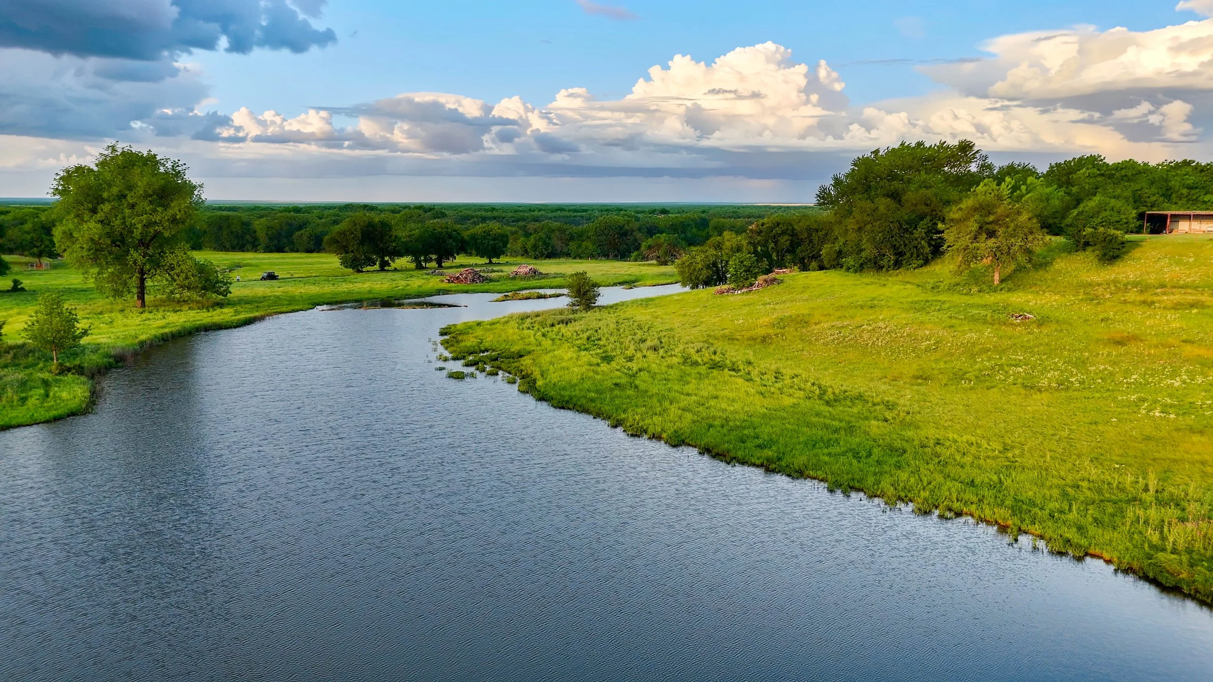 A scenic landscape featuring a river flowing through green grassy fields with trees, under a sky with scattered clouds.