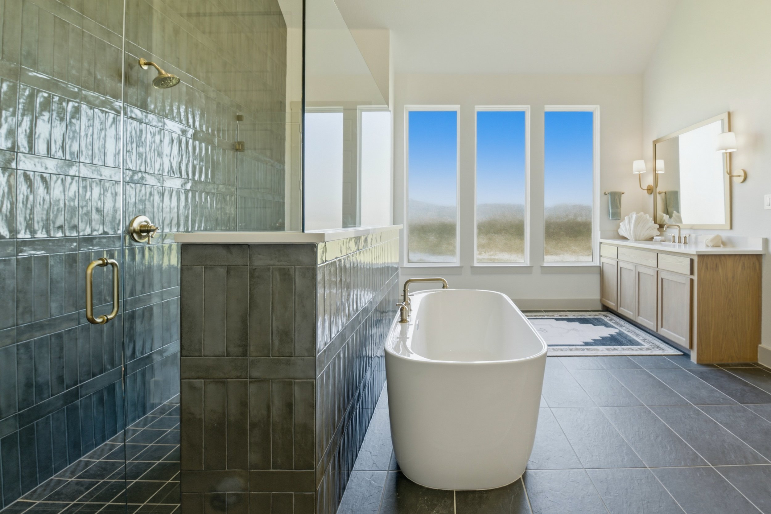 A modern bathroom with a freestanding bathtub in front of large windows showing a blue sky and landscape, a green-tiled shower area with brass fixtures, a wooden vanity with a large mirror, and minimal decor.