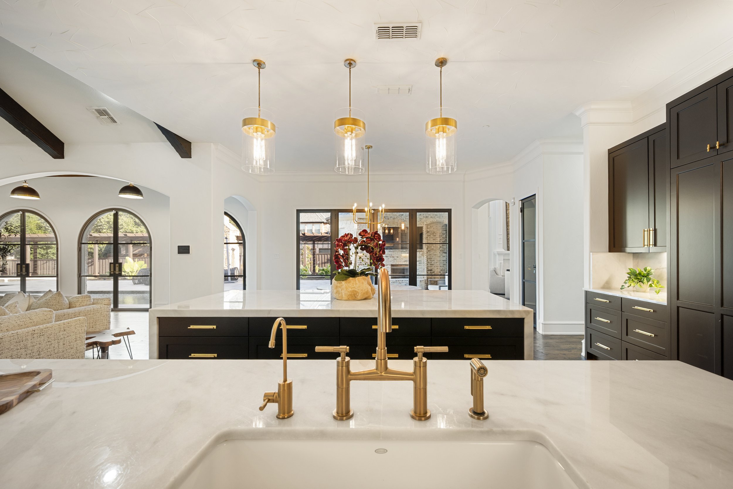 Modern kitchen with white marble island, gold fixtures, black cabinets, and a view of living space with arched windows and outdoor fencing.