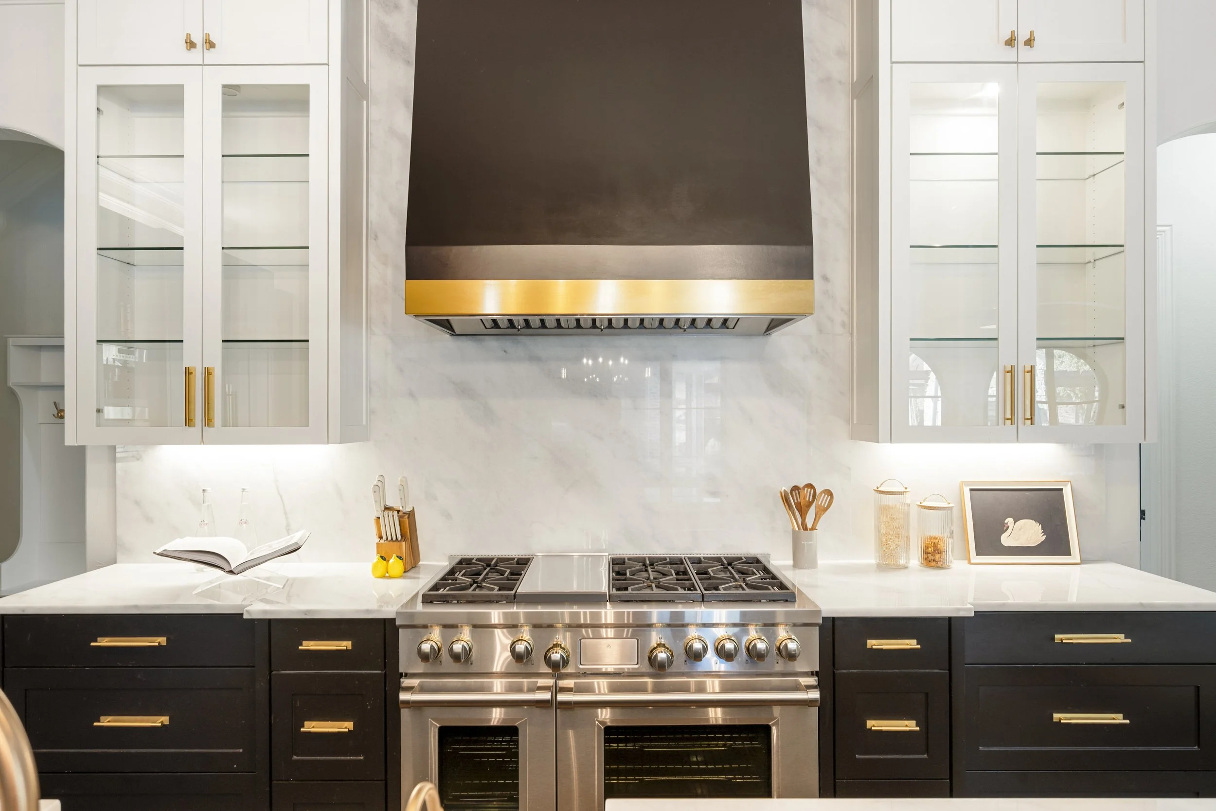 Modern kitchen with black cabinets and gold handles, stainless steel gas stove and oven, white marble countertops, open book, yellow decorative items, framed picture of a swan, utensils in a white jar, and glass canisters on the countertop.
