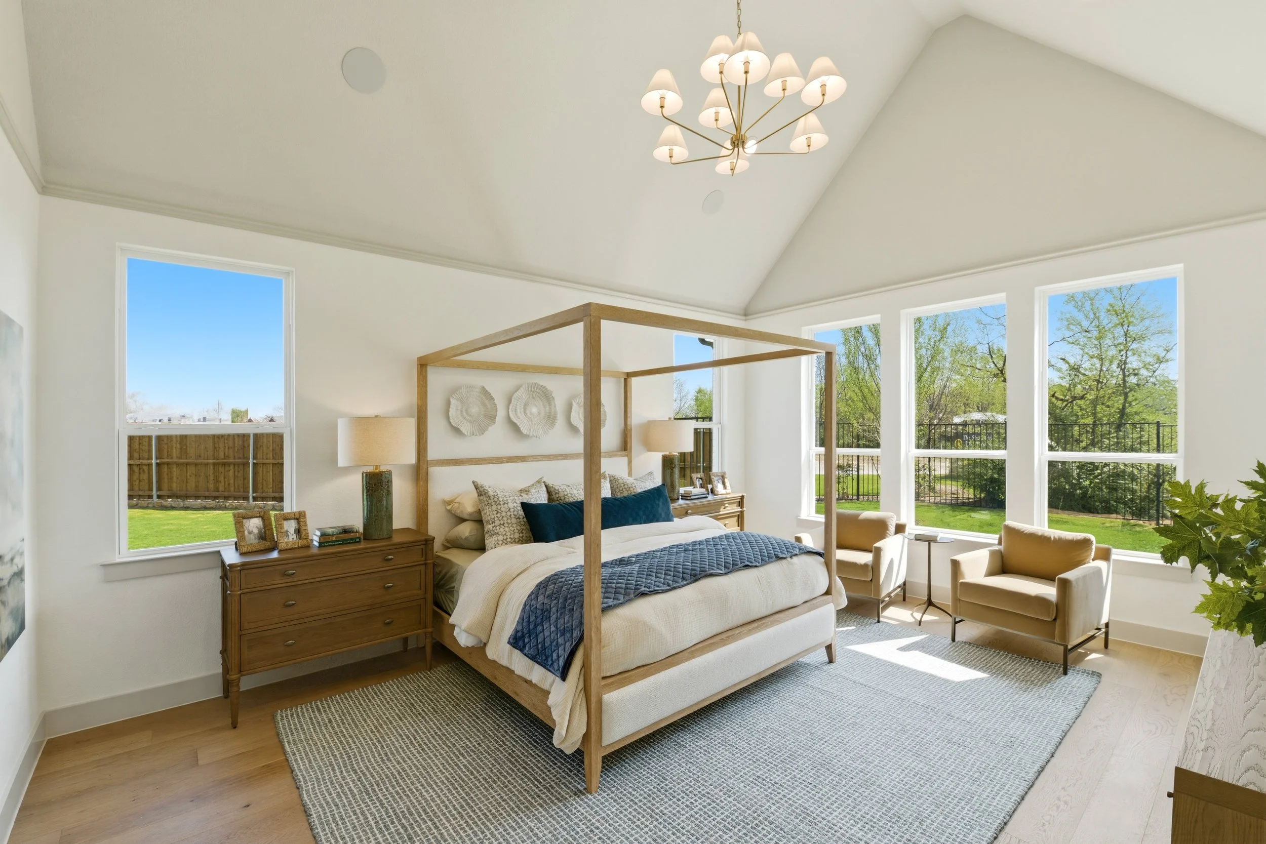 A bright bedroom with large windows and a view of green trees outside. It features a white bed with a wooden canopy, beige armchairs, a wooden dresser, and decorative wall art. Natural light fills the room, complemented by a modern chandelier.