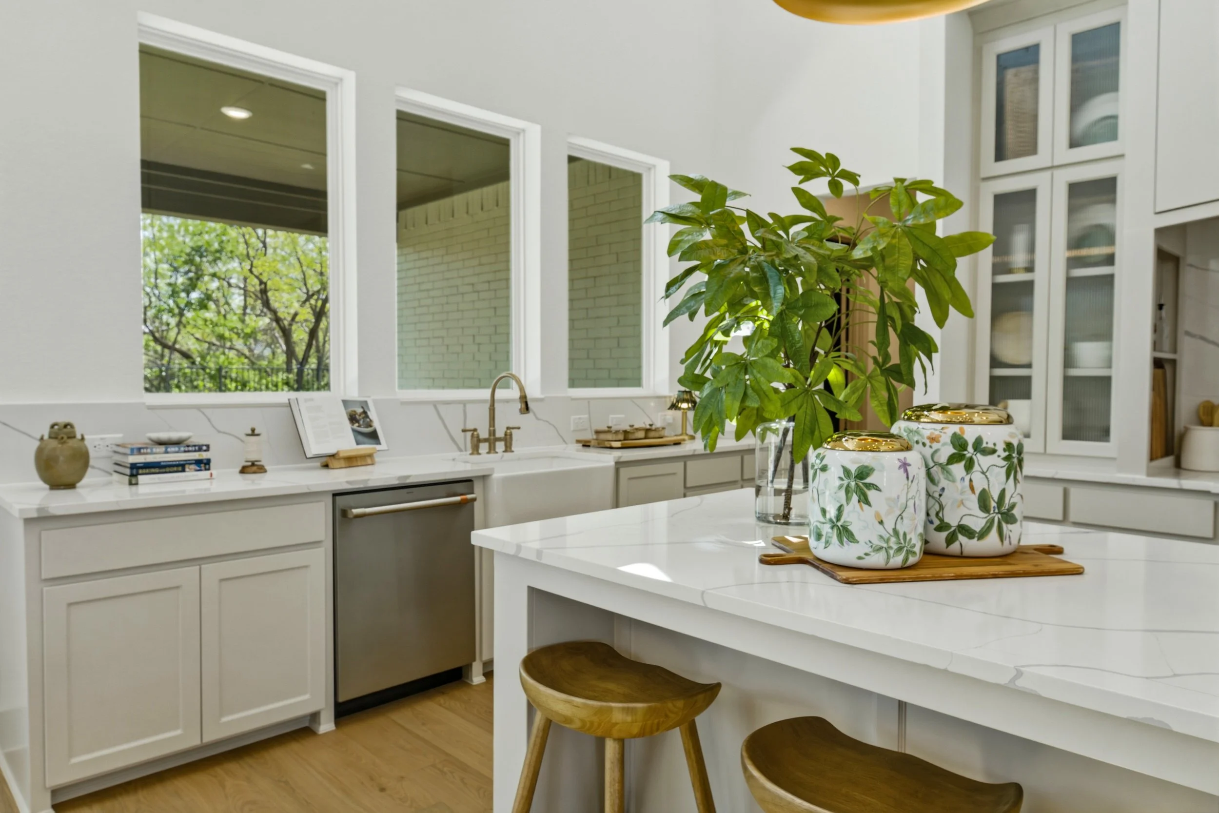 A modern kitchen with white cabinetry, a white marble island, large windows with greenery outside, a potted plant, decorative jars, and a few books on the counter.