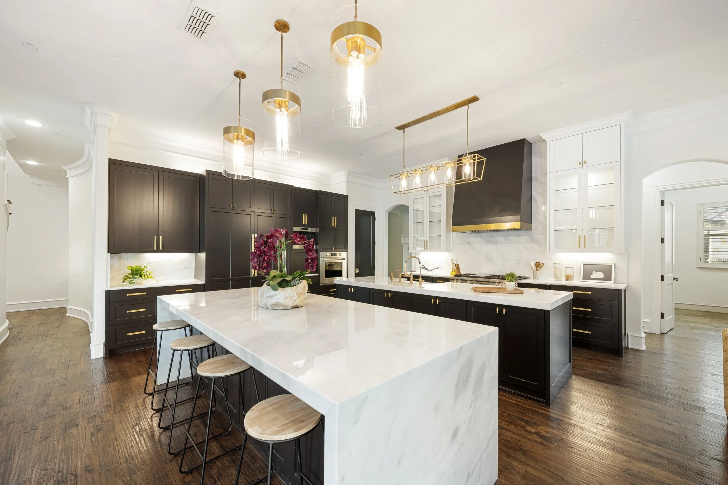Modern kitchen with dark cabinetry, white marble island, gold hardware, pendant lights, and hardwood floors. Pink orchids are on the island.